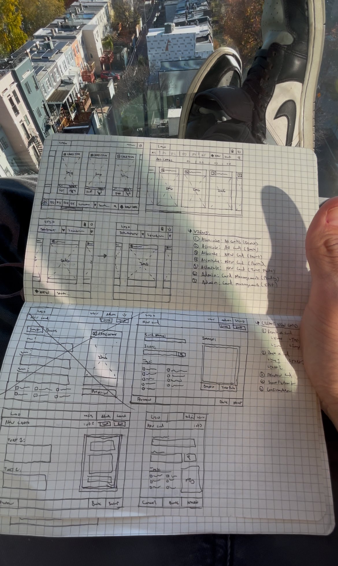 Open notebook with grid paper showing hand-drawn website or app wireframes, held by a person wearing black and white sneakers, with a cityscape view from above through a glass window.