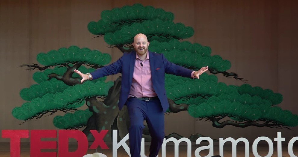 A man in a blue suit with a pink shirt standing in front of a painted tree background, with 'TEDxKumamoto' written at the bottom.