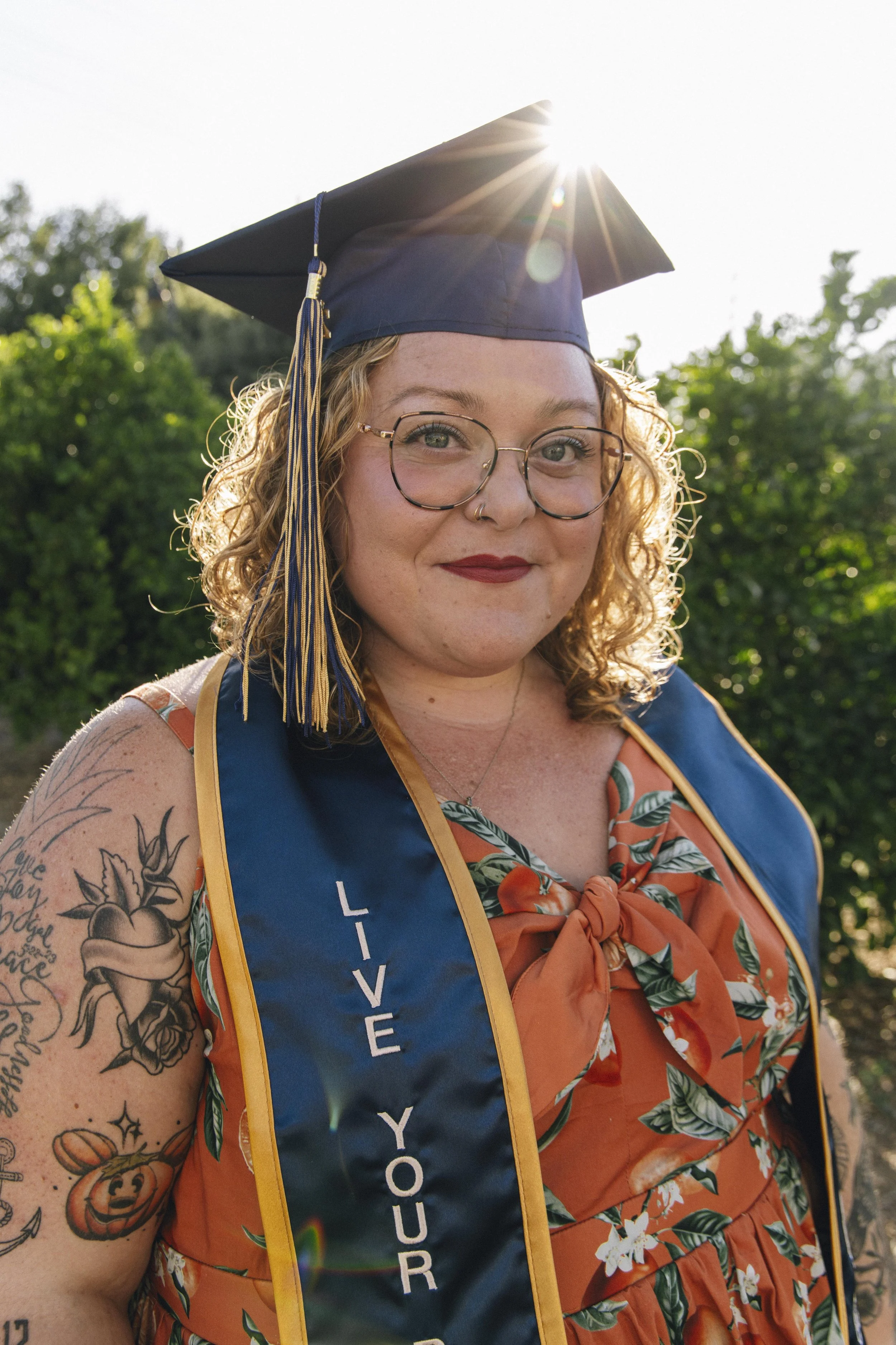A young woman with glasses and tattoos wearing a graduation cap and gown with a sash that says 'Live Your Purpose,' standing outdoors in the sunlight.