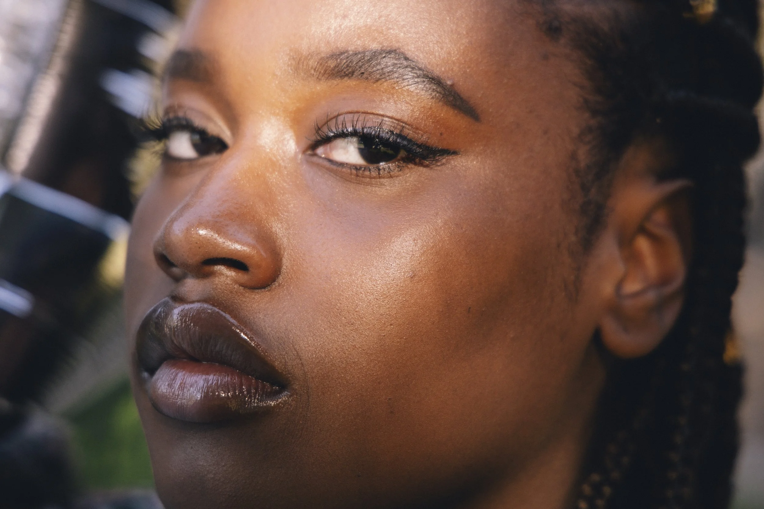 Close-up of a woman with dark skin, makeup, and braided hair, looking at the camera with one eye partially closed.