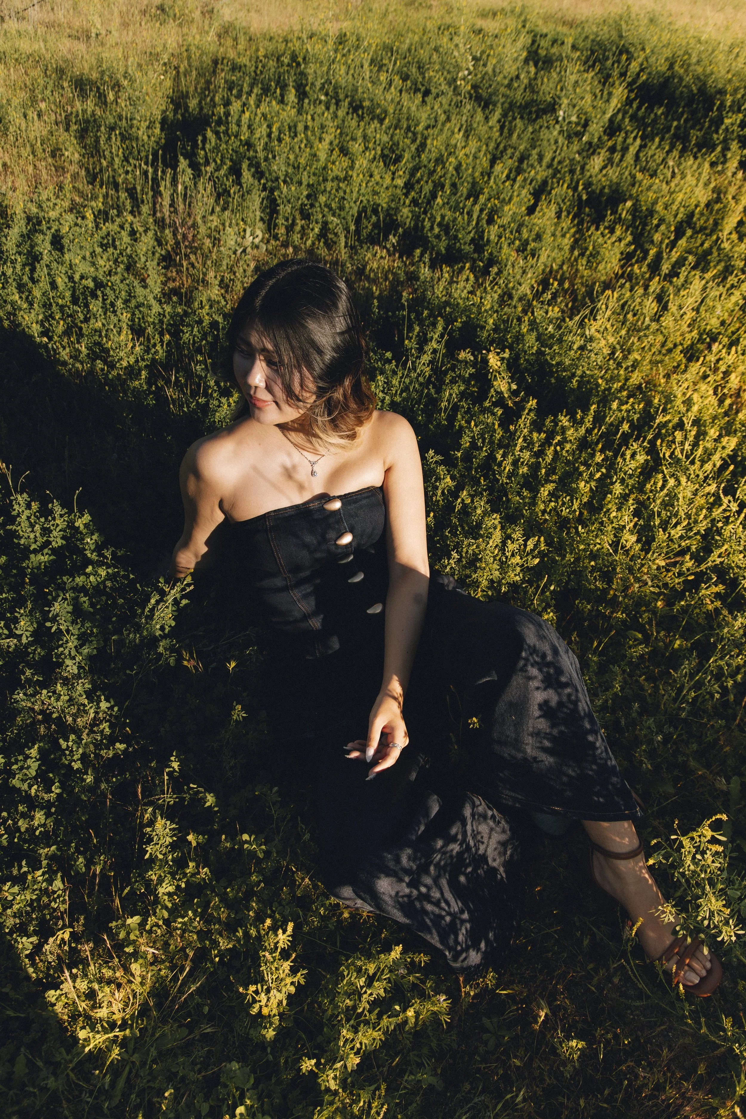 A woman sitting in a lush green field during sunset, wearing a black strapless dress with buttons down the front, and showing a necklace with a pendant.