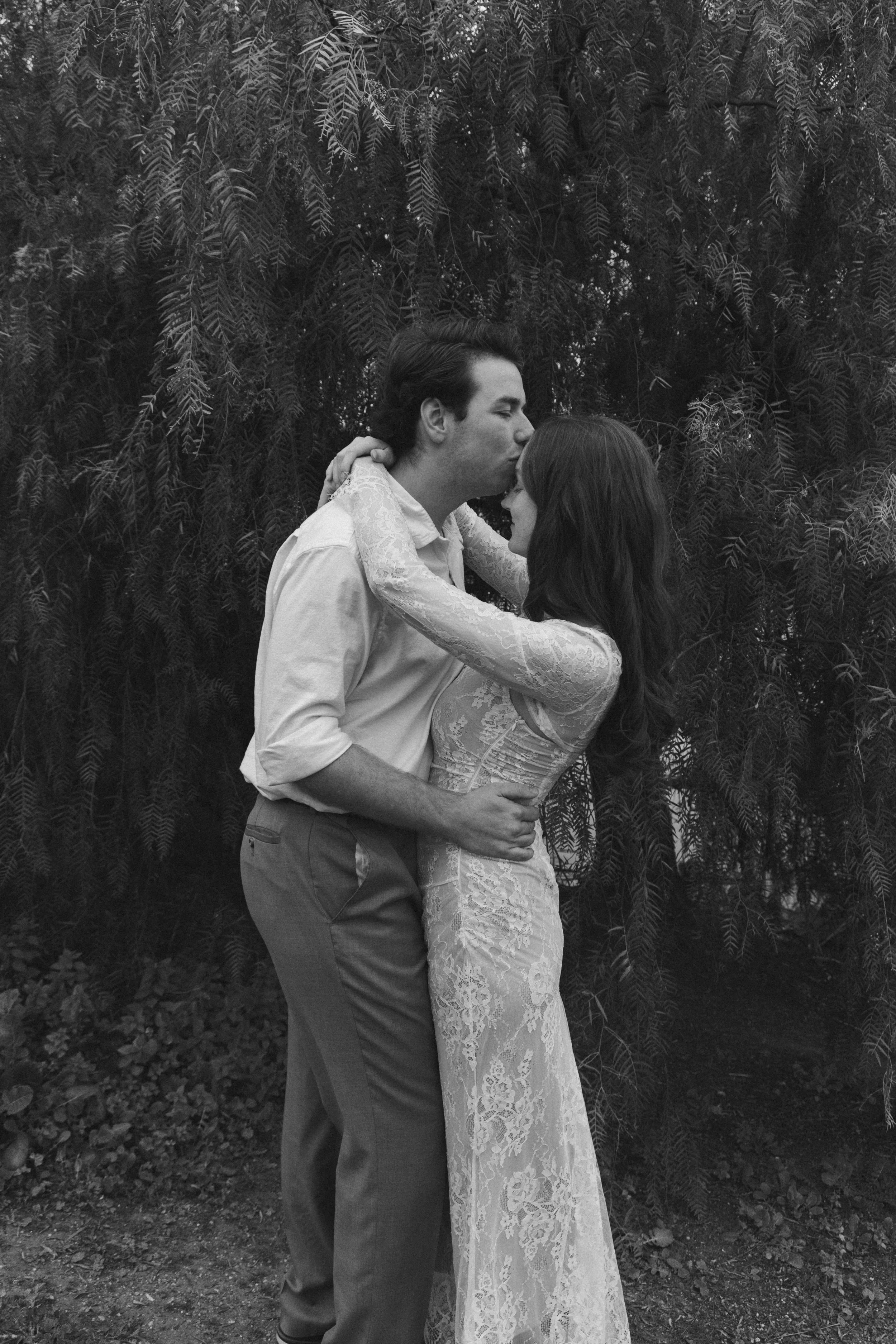 A black and white photograph of a couple embracing outdoors in front of a large tree with dense leaves. The man is kissing the woman's forehead while holding her close, and she is smiling with her eyes closed. The woman is wearing a lace dress and the man is dressed in a light-colored shirt and dark pants.
