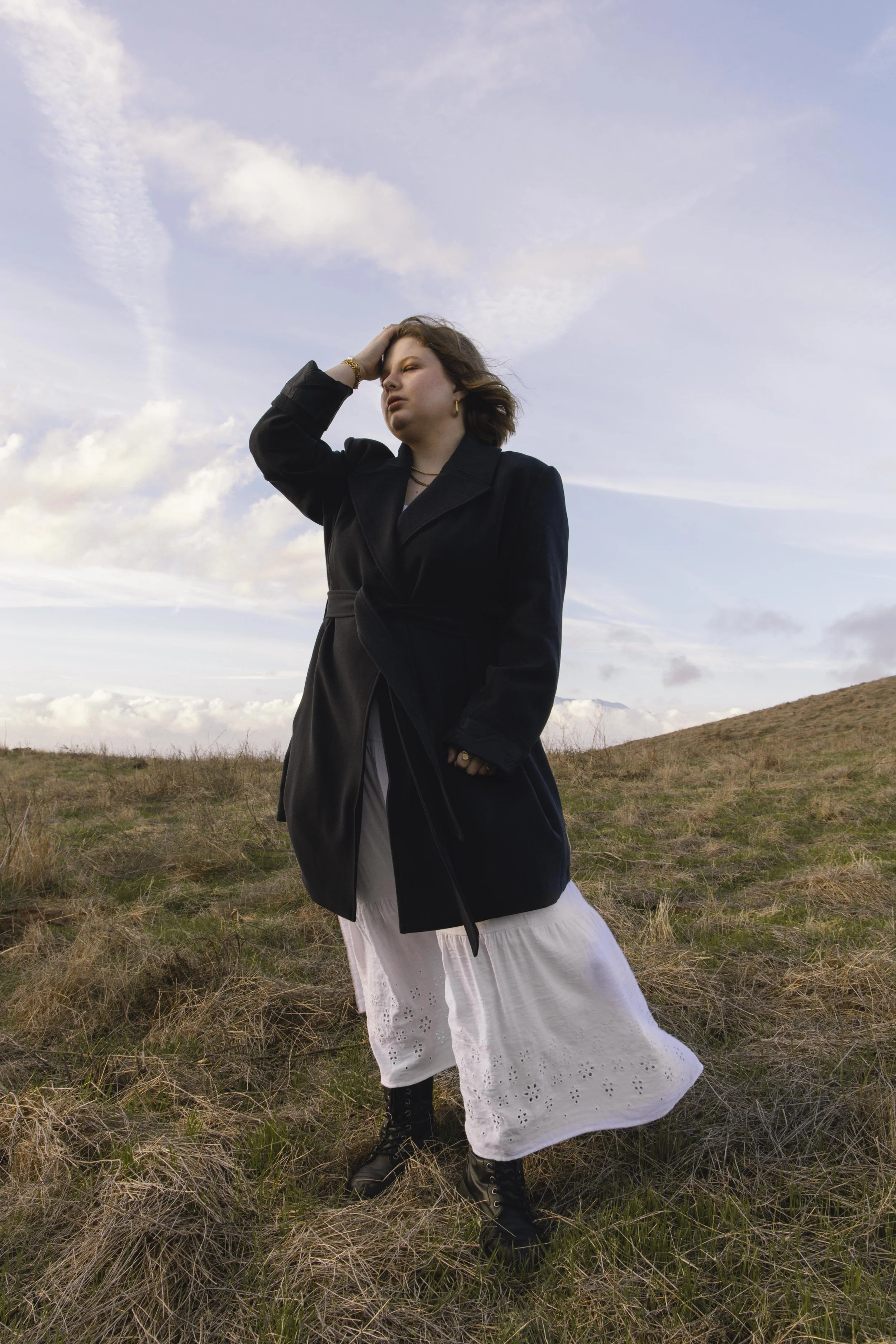 A woman standing in a grassy field with hills in the background, wearing a black coat, a white skirt, black boots, and gold jewelry, holding her head with one hand under a cloudy sky.