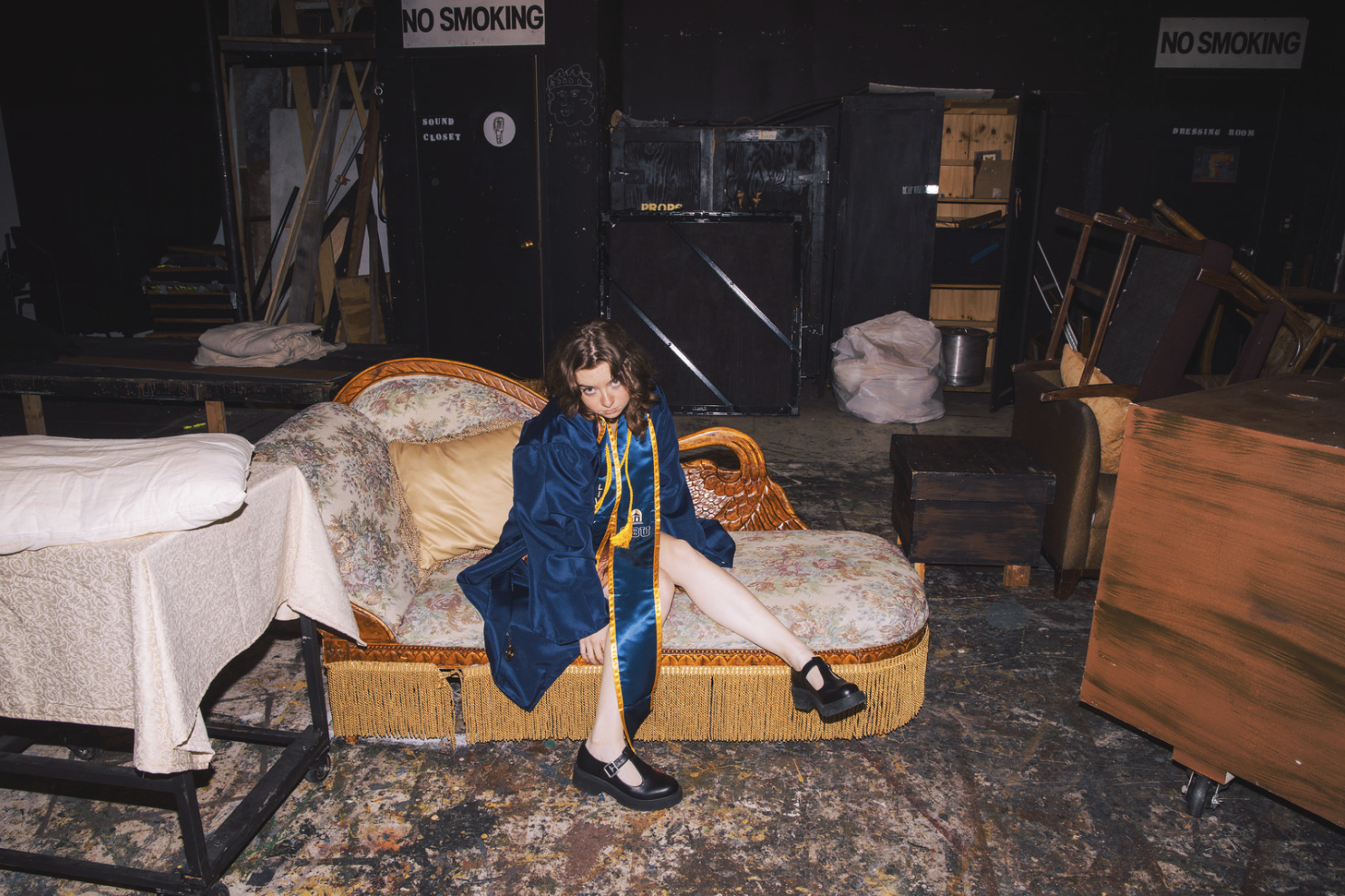 Young woman with curly brown hair sitting on an ornate floral sofa in a cluttered backstage or storage area, wearing a blue graduation gown and black shoes, looking at the camera.