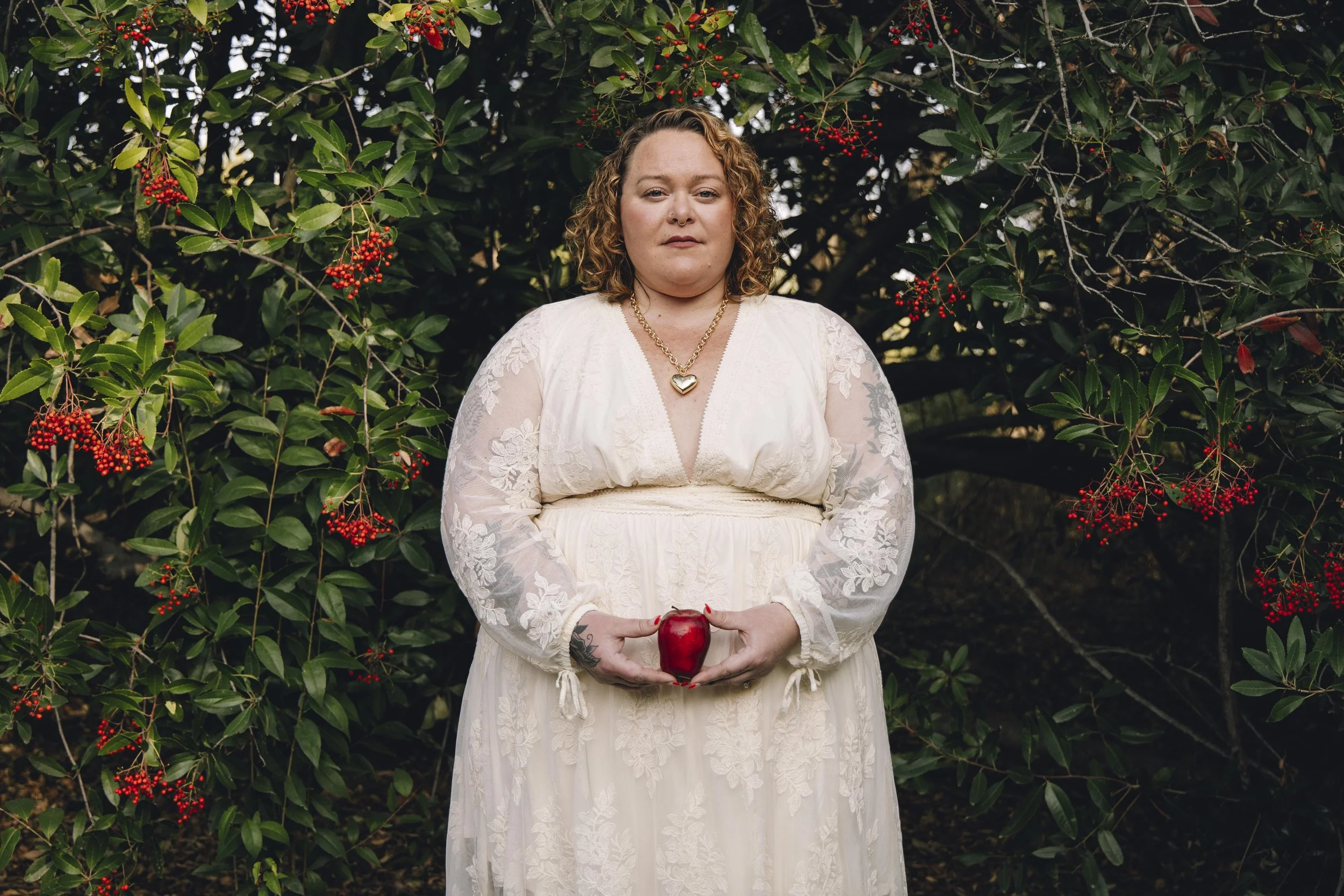 A woman with curly hair, wearing a white lace dress and a gold heart necklace, stands in front of a bush with red berries, holding a red apple in her hands.
