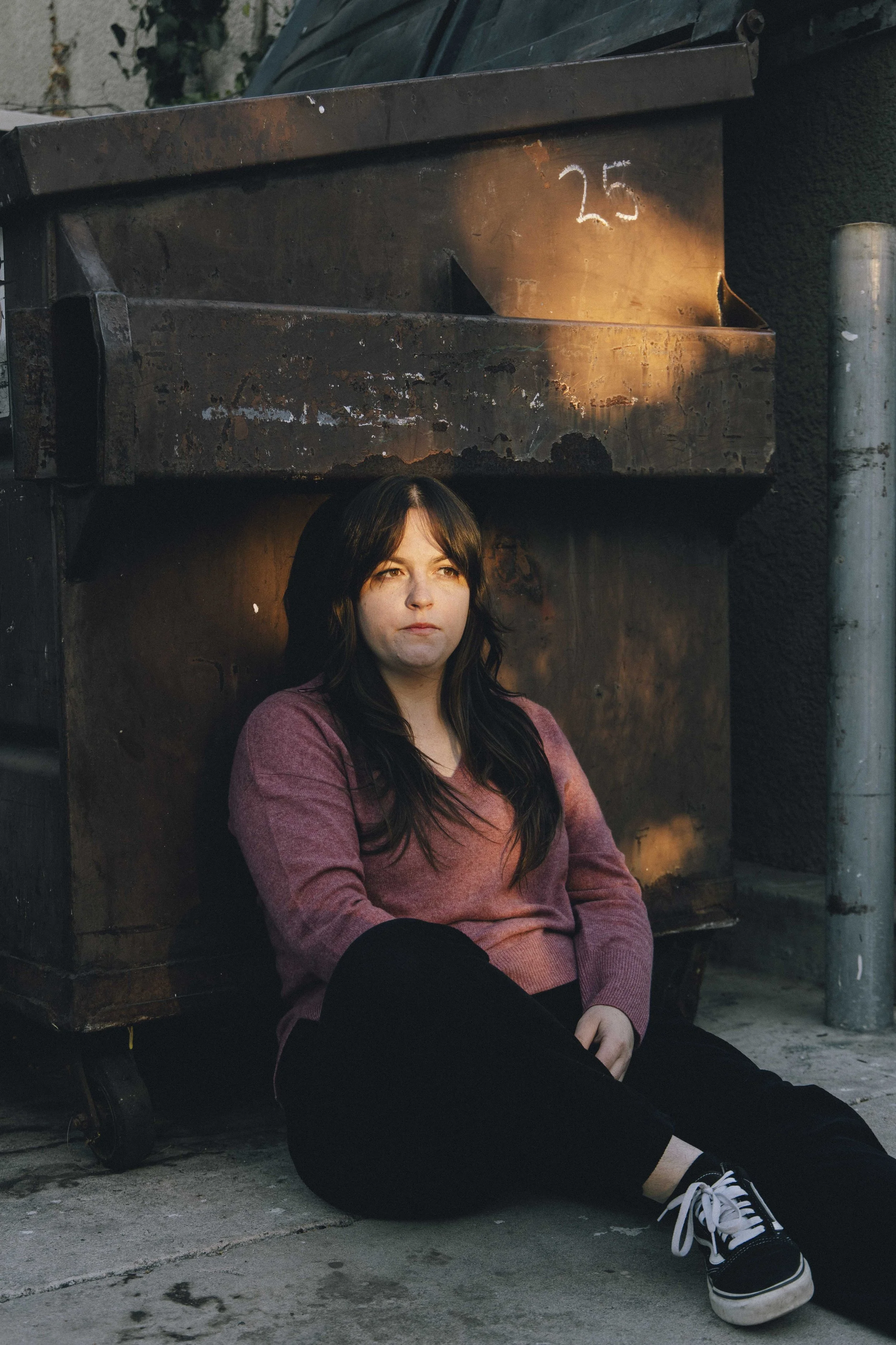 A young woman with long dark hair sitting on the ground in front of a rusted metal dumpster. She is wearing a pinkish-purple long-sleeve shirt, black pants, and black sneakers with white soles and laces. The dumpster has the number 25 written on it i