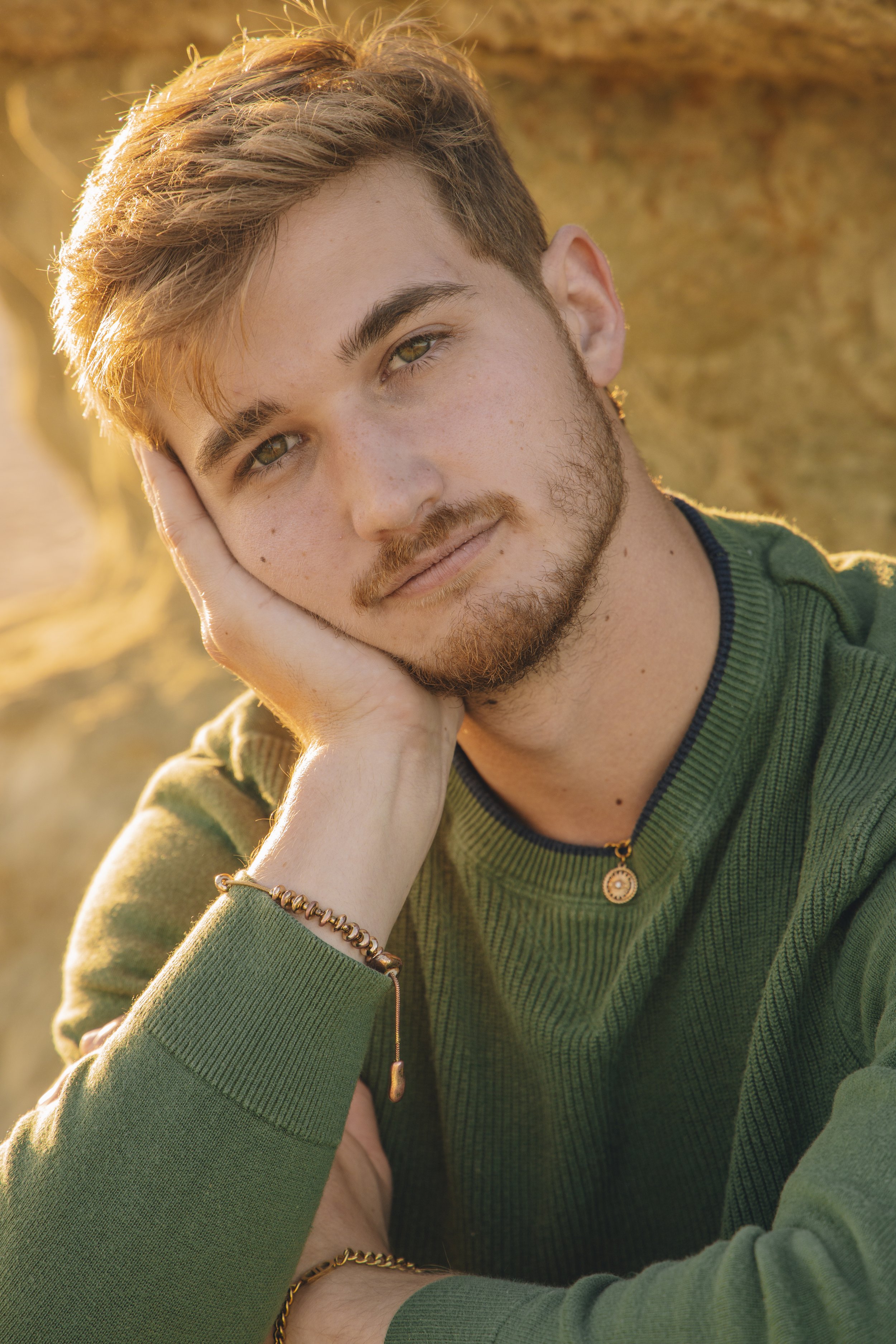 A young man with light brown hair and hazel eyes is resting his face on his hand while smiling softly. He is wearing a green sweater, a gold necklace, and a bracelet on his wrist. The background is an outdoor scene with warm, golden lighting, likely 