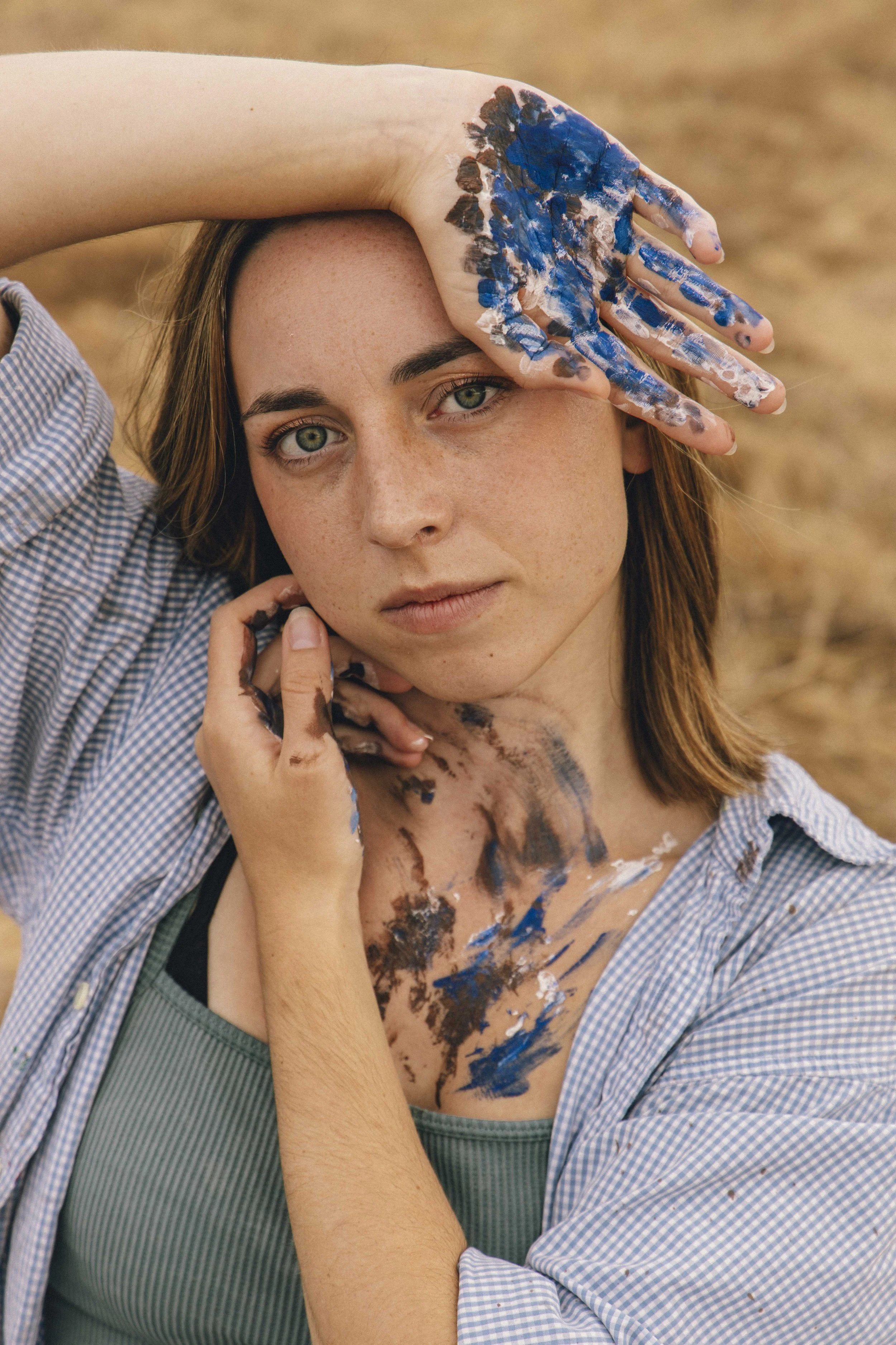 A woman with light brown hair, blue eyes, and freckles looks directly at the camera while her face and arm are covered in blue, white, and brown paint. She is wearing a blue and white checkered shirt and a green top underneath.