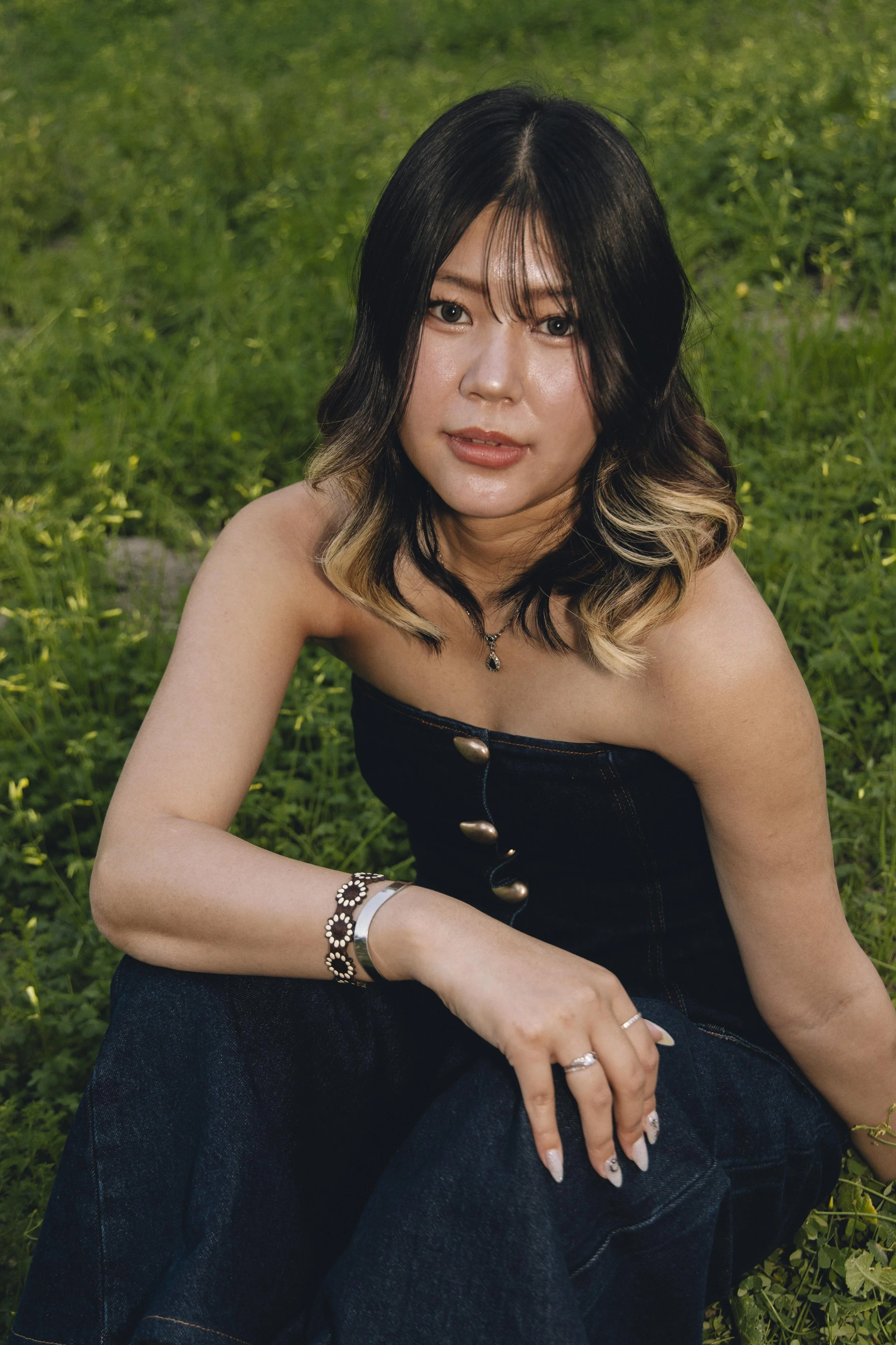 A woman with long black hair with blonde tips, wearing a black strapless top, jewelry, and dark jeans, sitting outdoors on green grass.