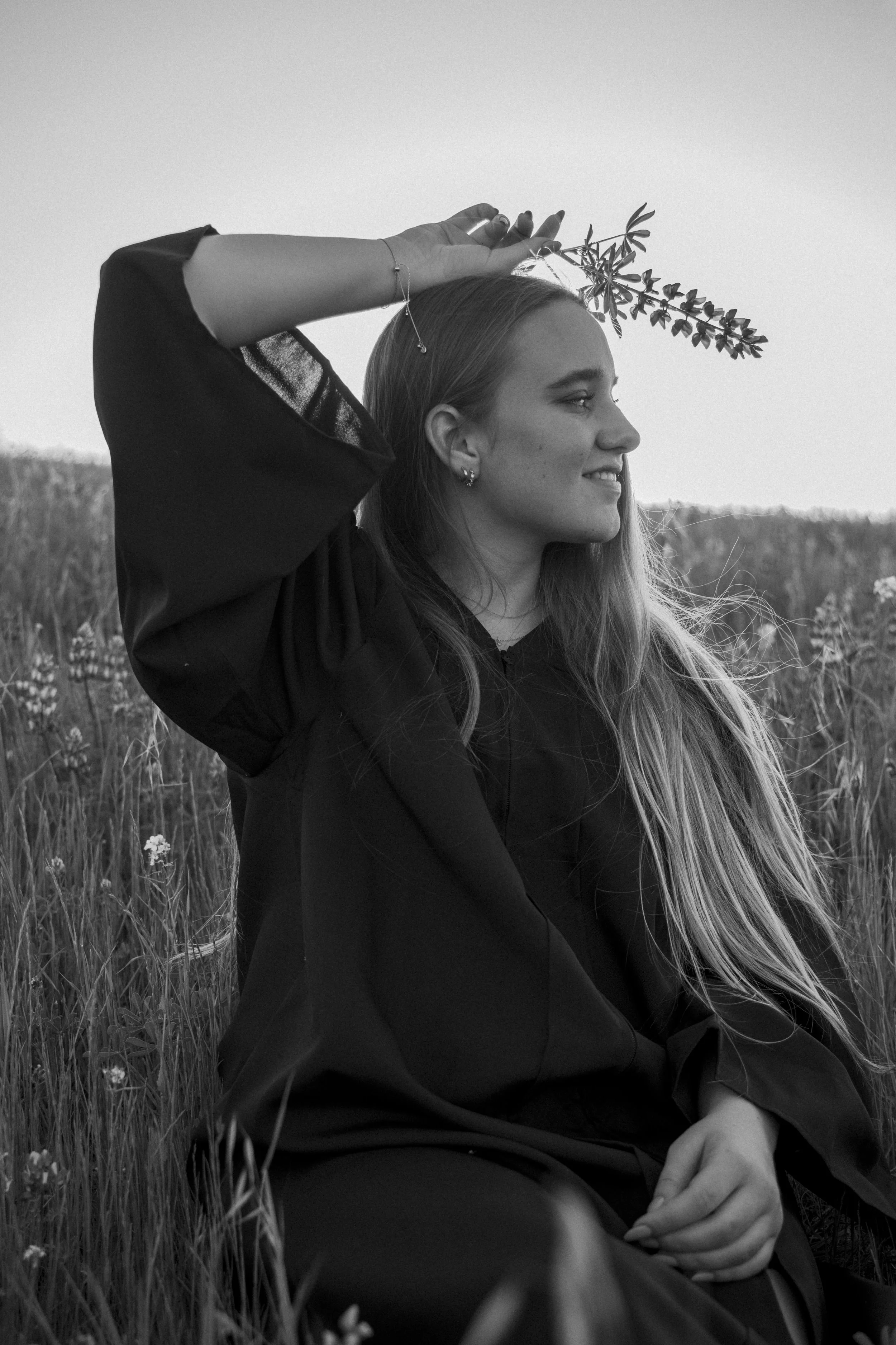 A woman sitting in a field holding a small flower stem on her head, smiling and looking to the side in the California hills.