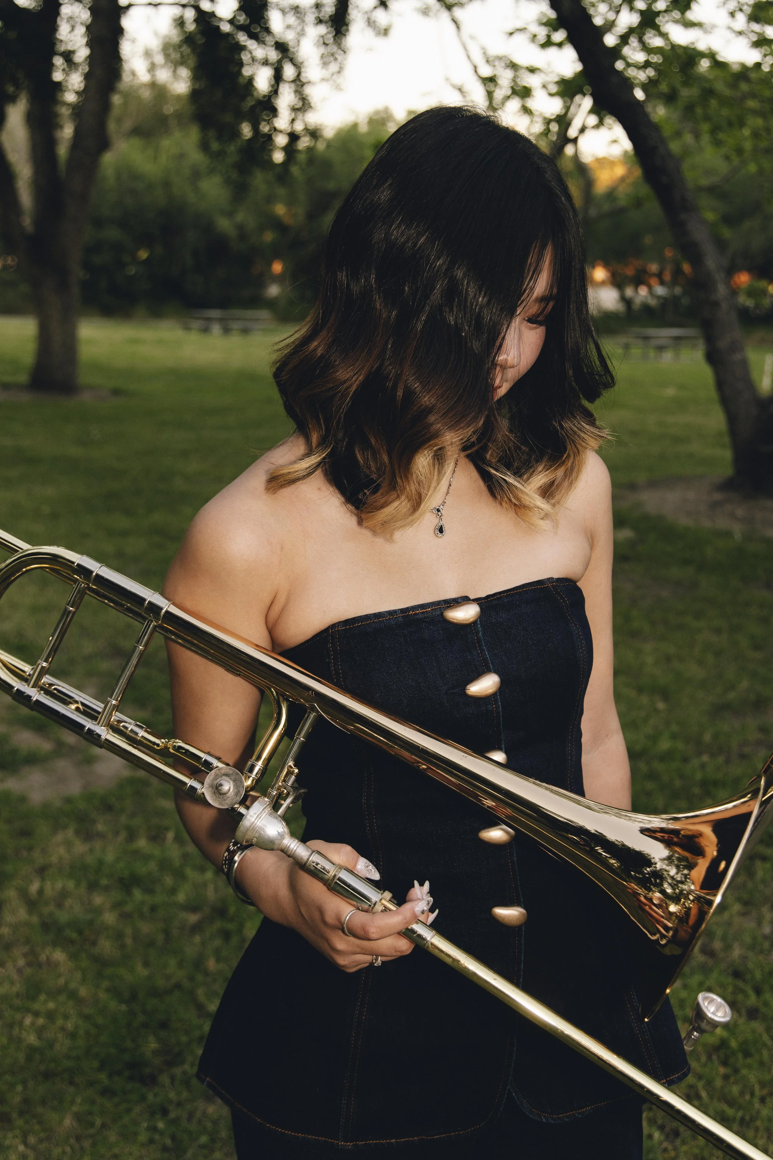 A young woman with shoulder-length black hair with blonde tips, wearing a strapless denim dress with large buttons, holds a shiny brass saxophone and stands outdoors in a park with trees in the background.