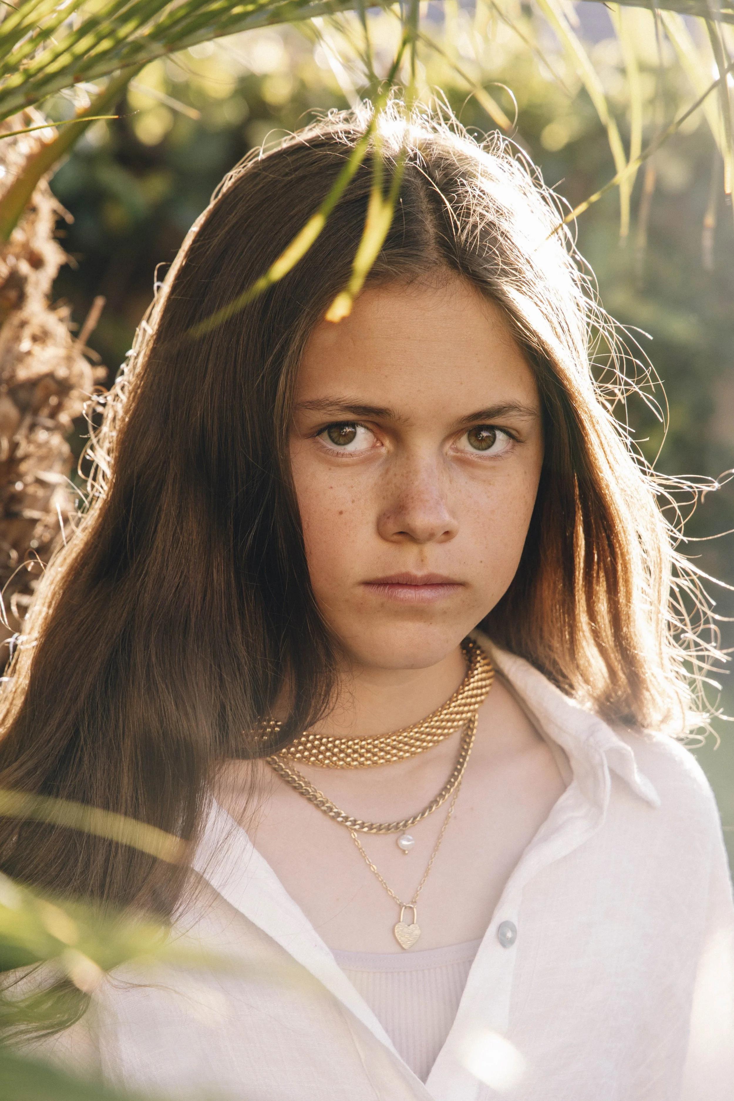Close-up of a young woman with long brown hair, wearing layered gold necklaces and a white button-up shirt, standing outdoors with sunlight backlighting her face, partially obscured by green leaves.
