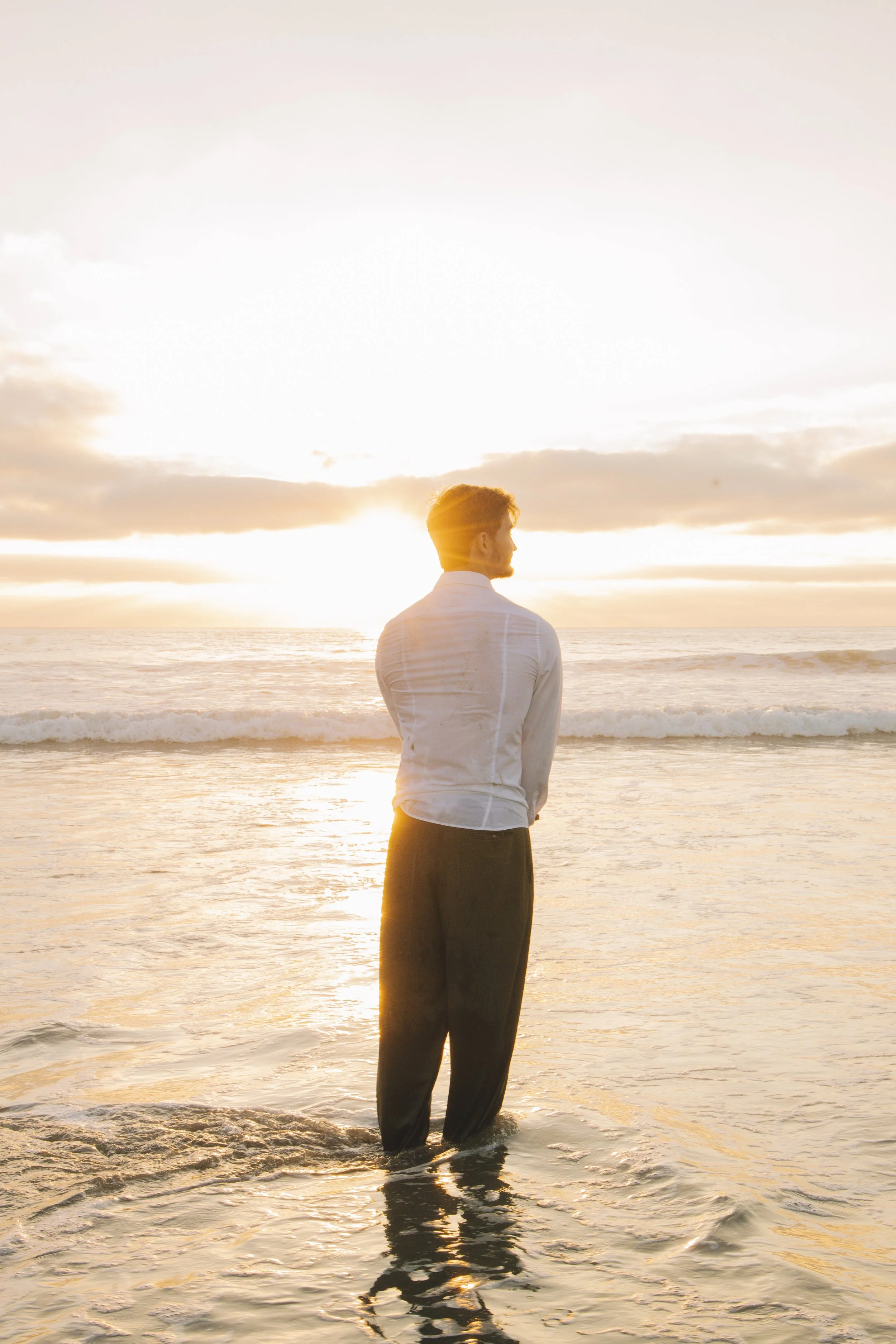 A man dressed in formal clothes standing on the beach at sunset, facing the horizon and the setting sun.