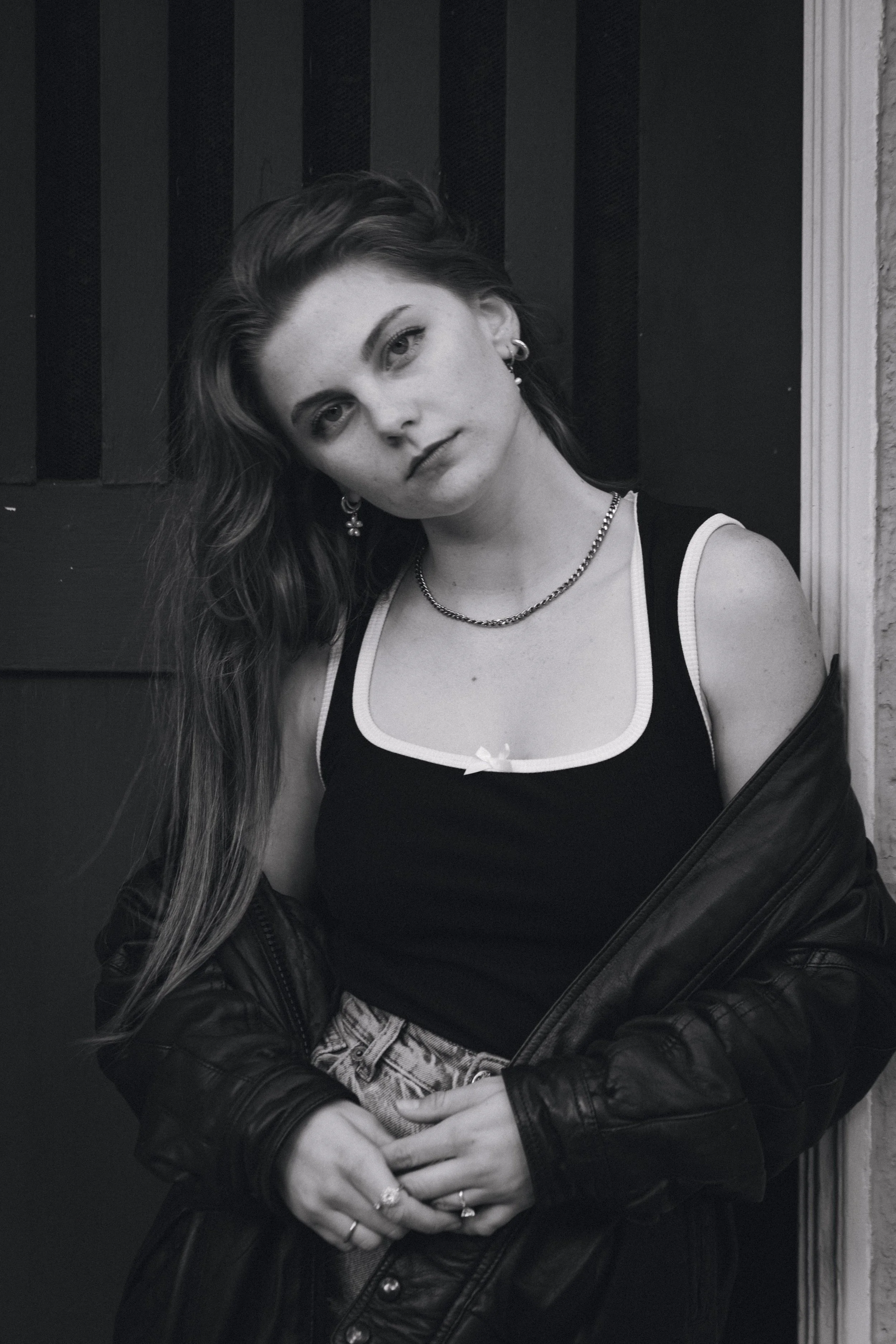 A young woman with long wavy hair, wearing earrings, a necklace, a black tank top with white trim, a leather jacket hanging off her shoulders, and patterned pants, posing against a dark background with a serious expression.