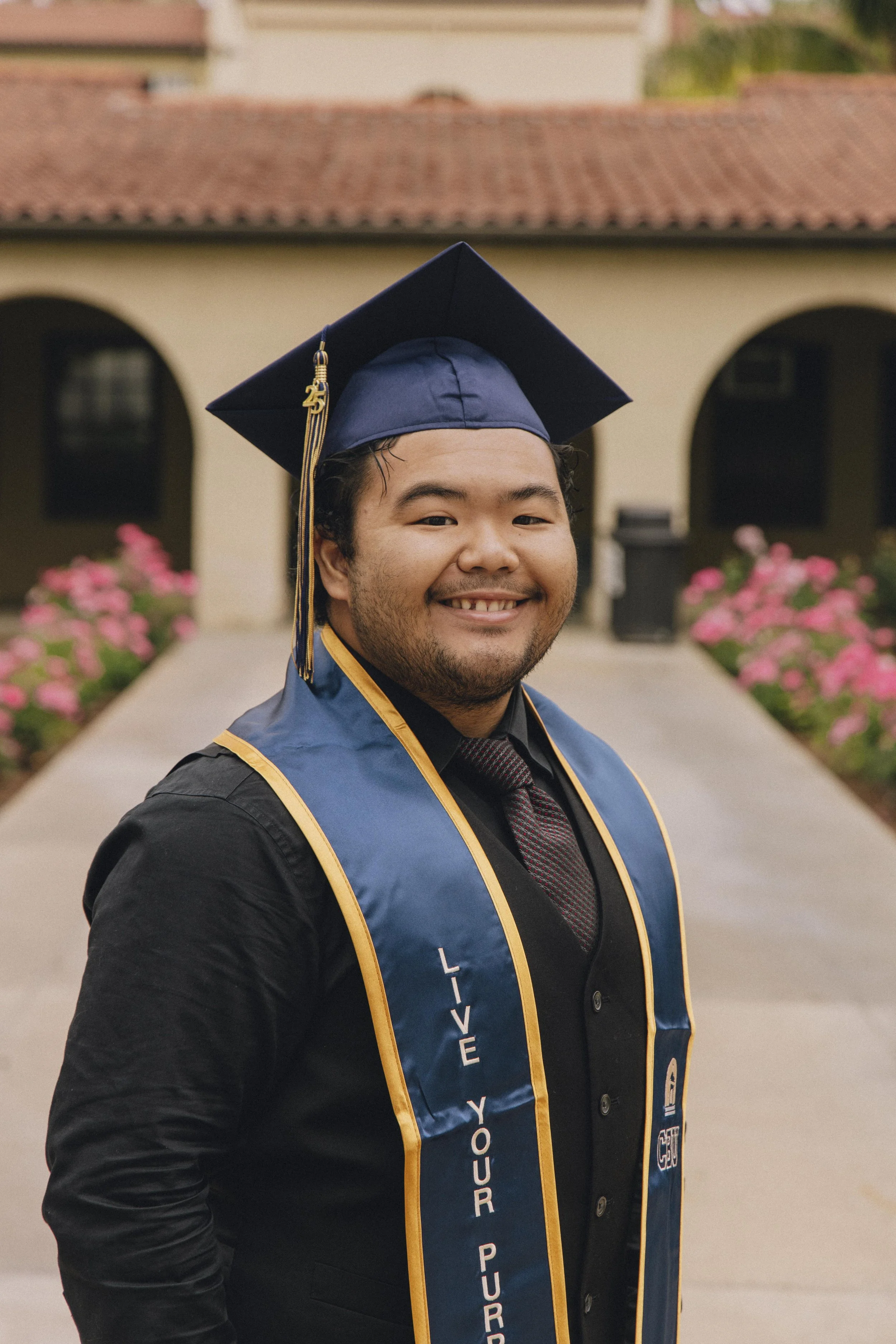 A smiling man in a graduation cap and gown stands outdoors on a walkway with pink flowers on either side, in front of a building with arches and a tiled roof, celebrating graduation.