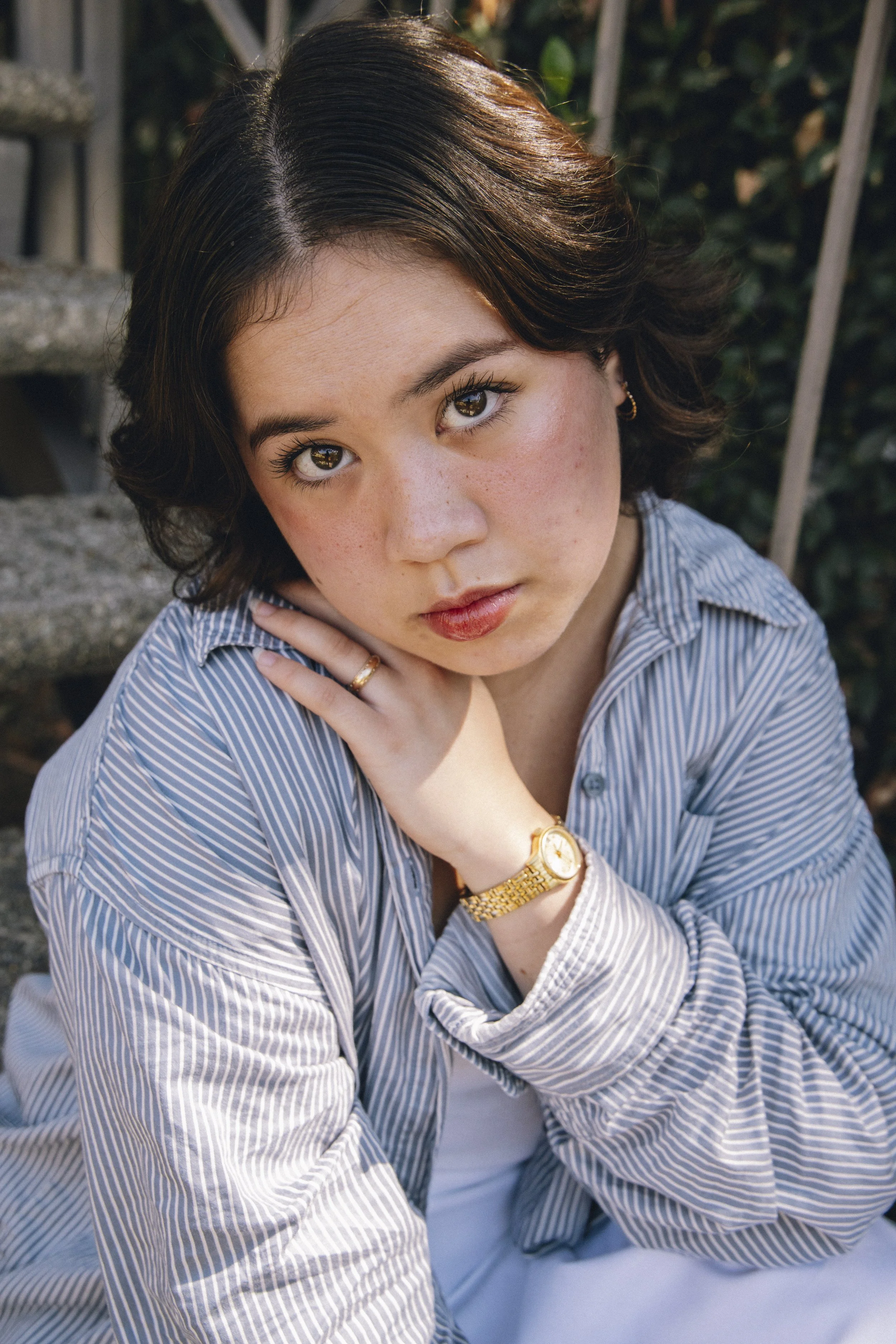 A young woman with brown hair and brown eyes, wearing a striped shirt, a gold watch, and rings, sitting outdoors near a staircase and green plants, looking directly at the camera.