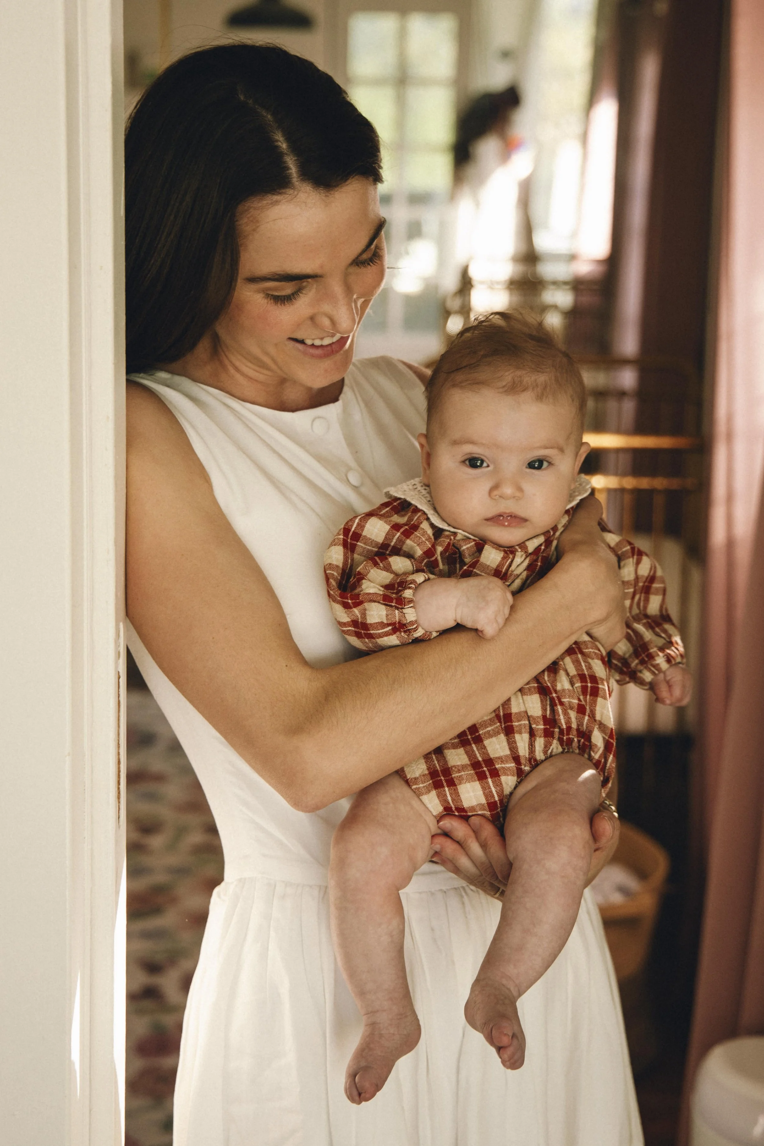 A woman with dark hair, smiling, holding a baby with light hair, dressed in a red and beige checkered outfit, in a warmly lit room.