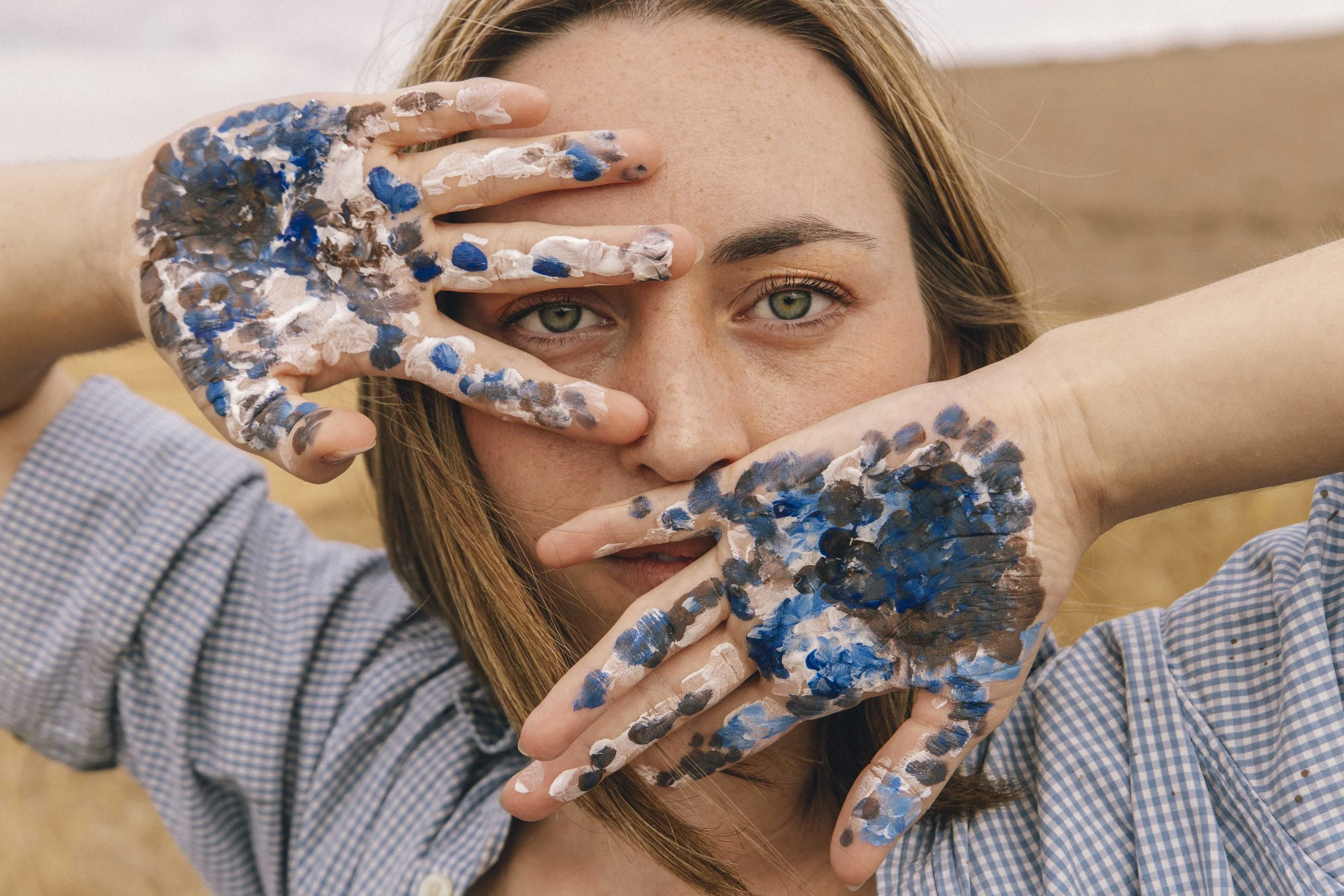 A woman with light skin and green eyes, holding her face with multiple hands covered in blue, white, black, and brown paint, partially covering her face, with a blurred outdoor background.