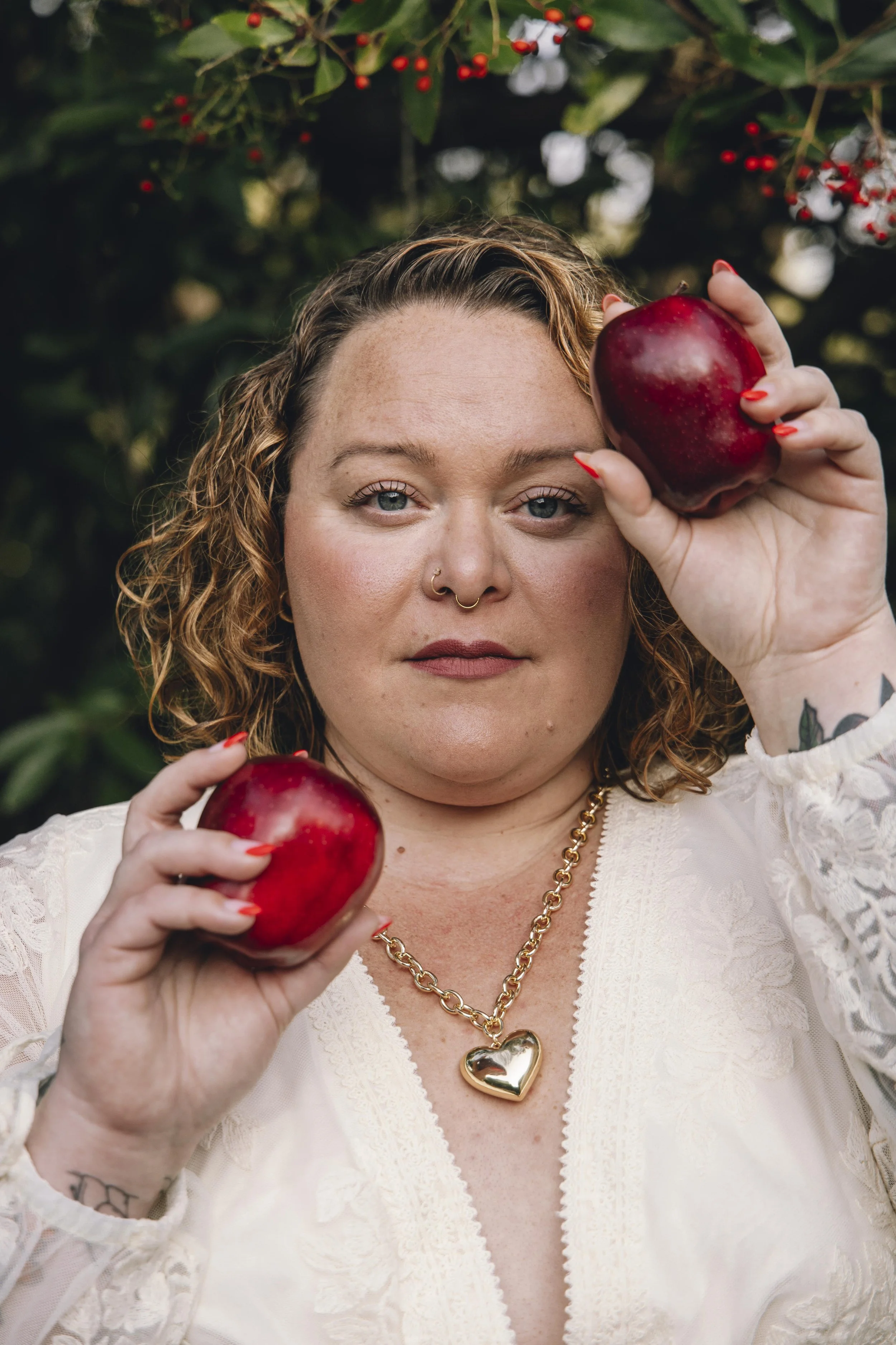 A woman with curly hair and a nose ring holds two large red apples, one close to her face and the other in her hand, standing outdoors with green foliage and red berries in the background.