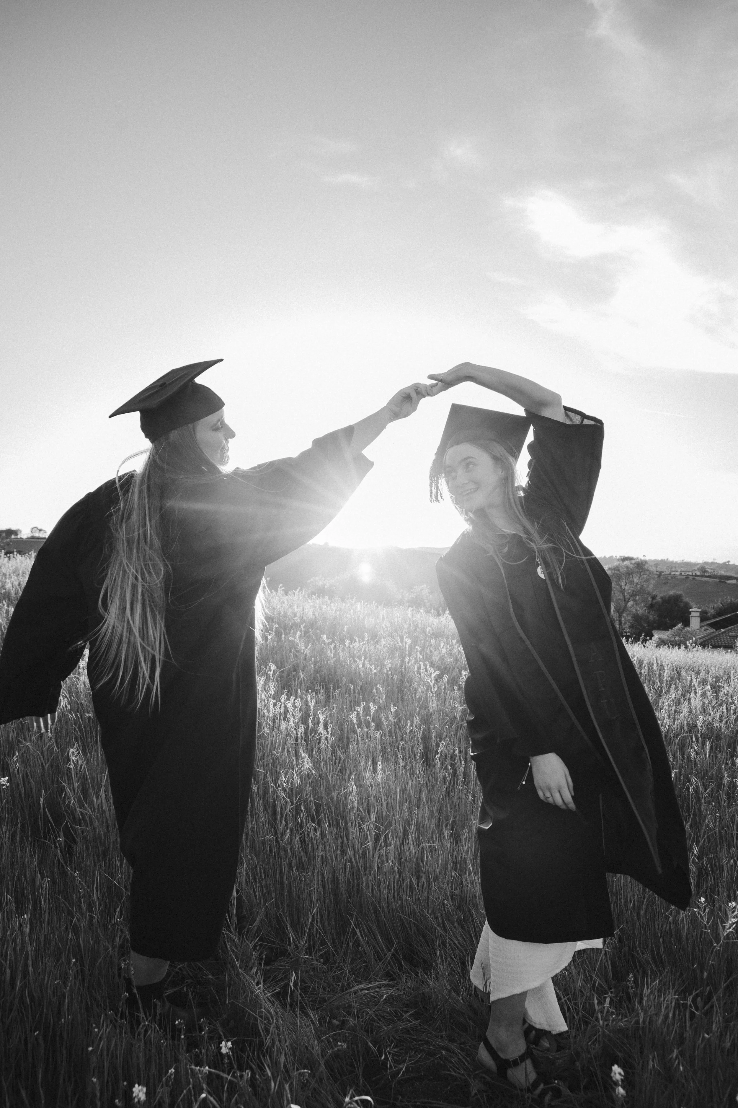 Two young women in graduation caps and gowns exchange a high five outdoors at sunset, with a grassy field and hills in the California foothills.