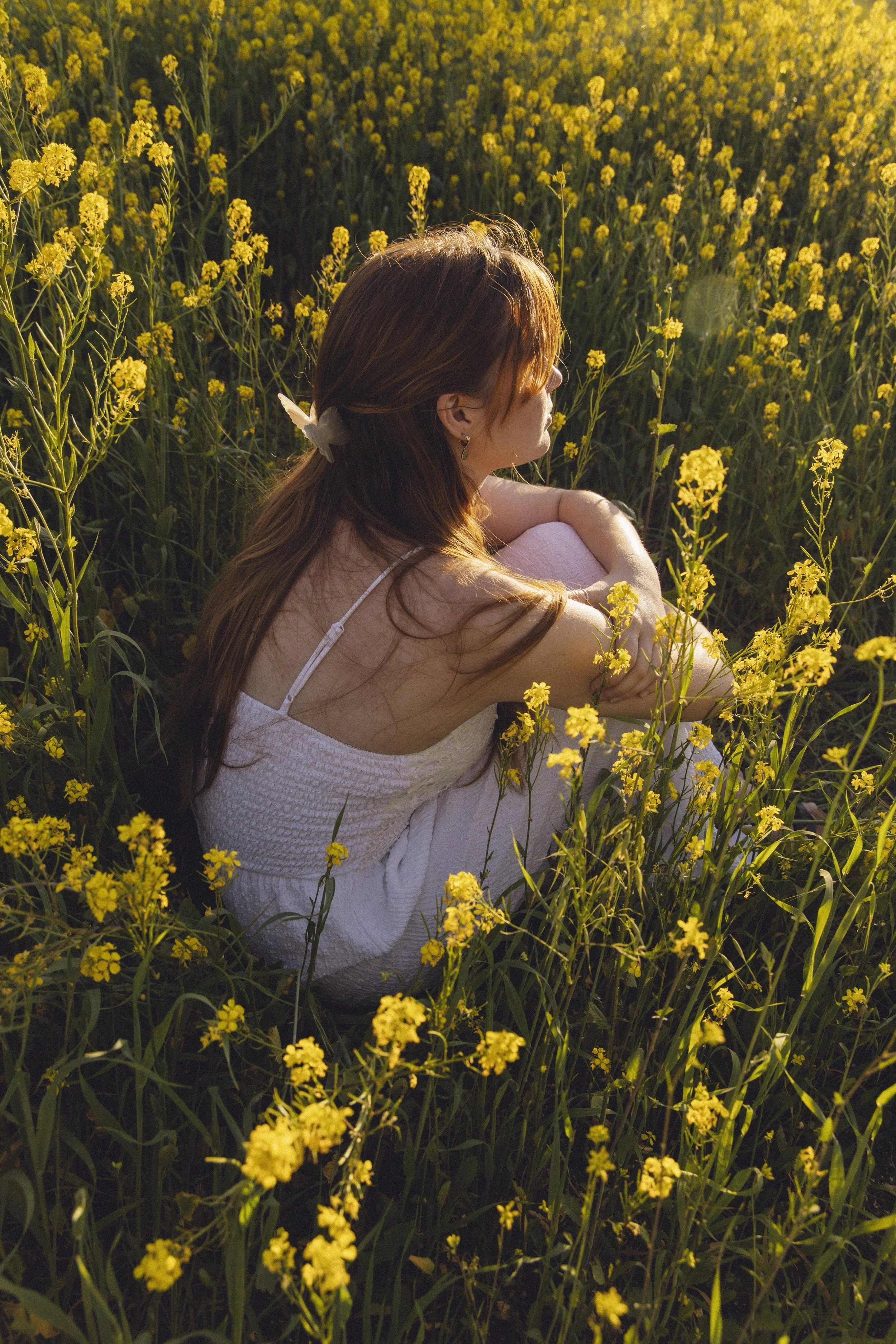 A woman with long brown hair sitting in a field of tall yellow flowers, facing sideways with her eyes closed, during sunset.