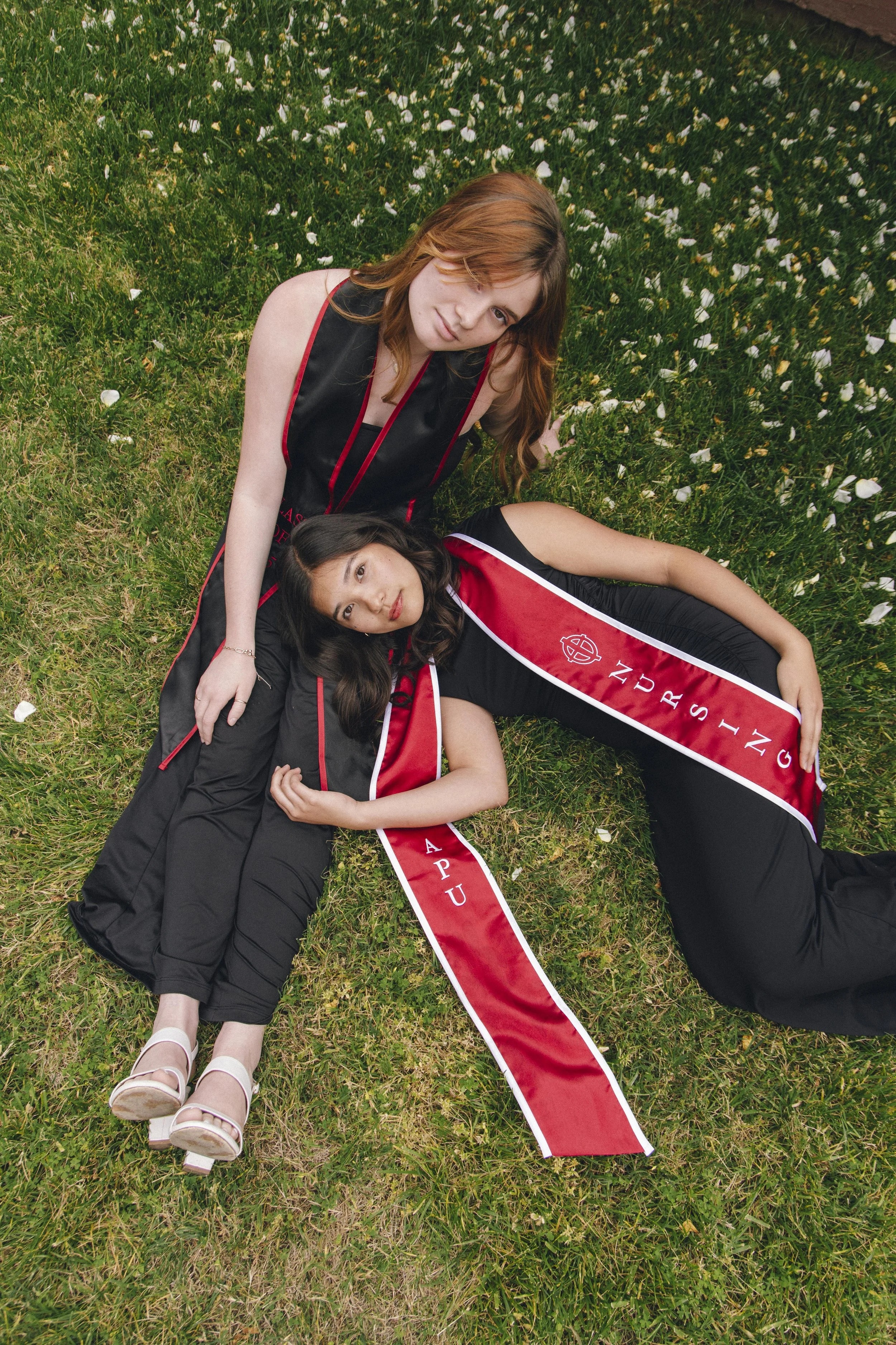 Two young women wearing black graduation gowns with red and white stoles lying on a grassy field with white flower petals, one with red hair and the other with dark hair and medium skin, posing for the photo.