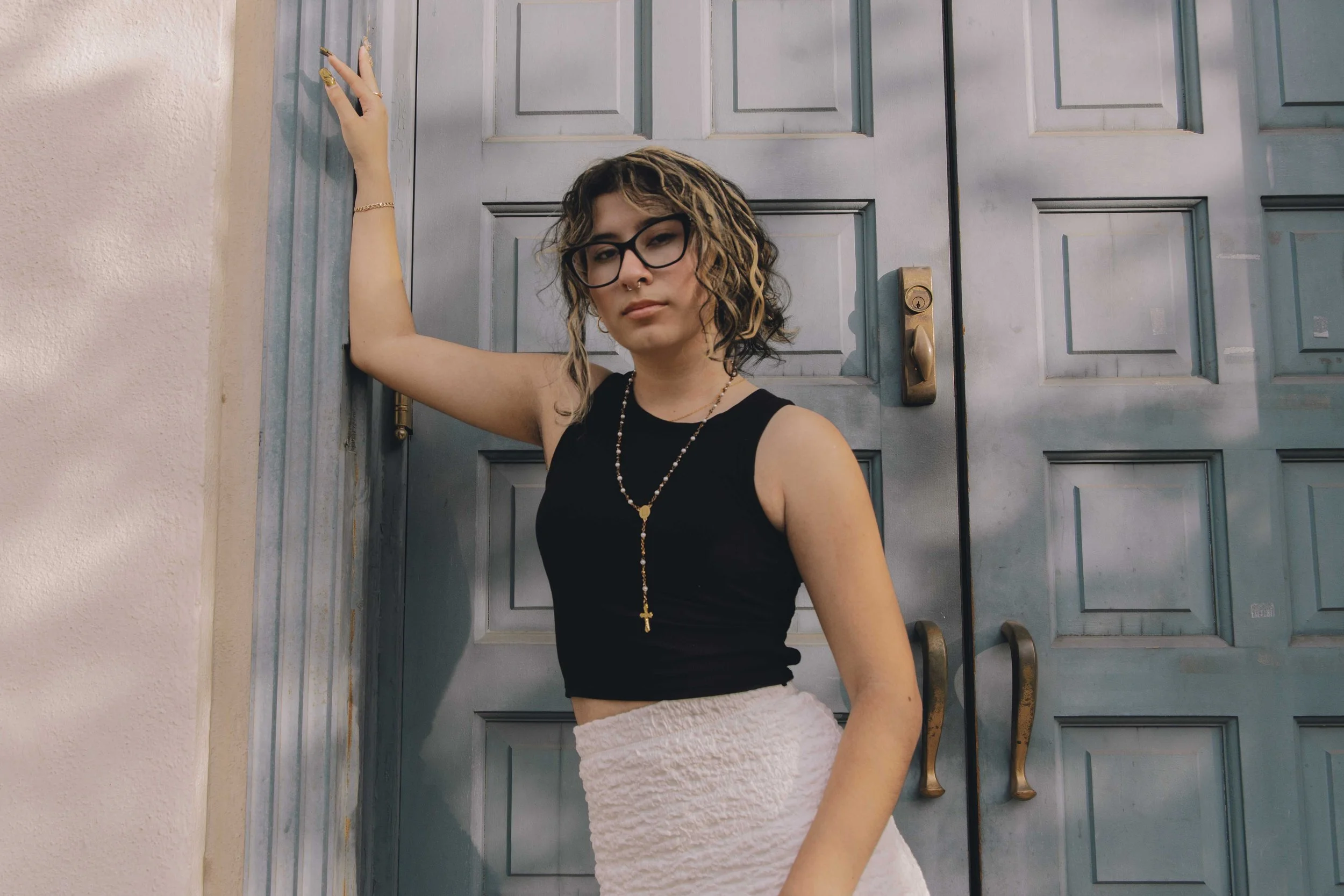 A young woman with curly hair, glasses, and piercings, wearing a black sleeveless top and a long necklace, standing in front of a blue door with her arm raised and hand touching the wall.