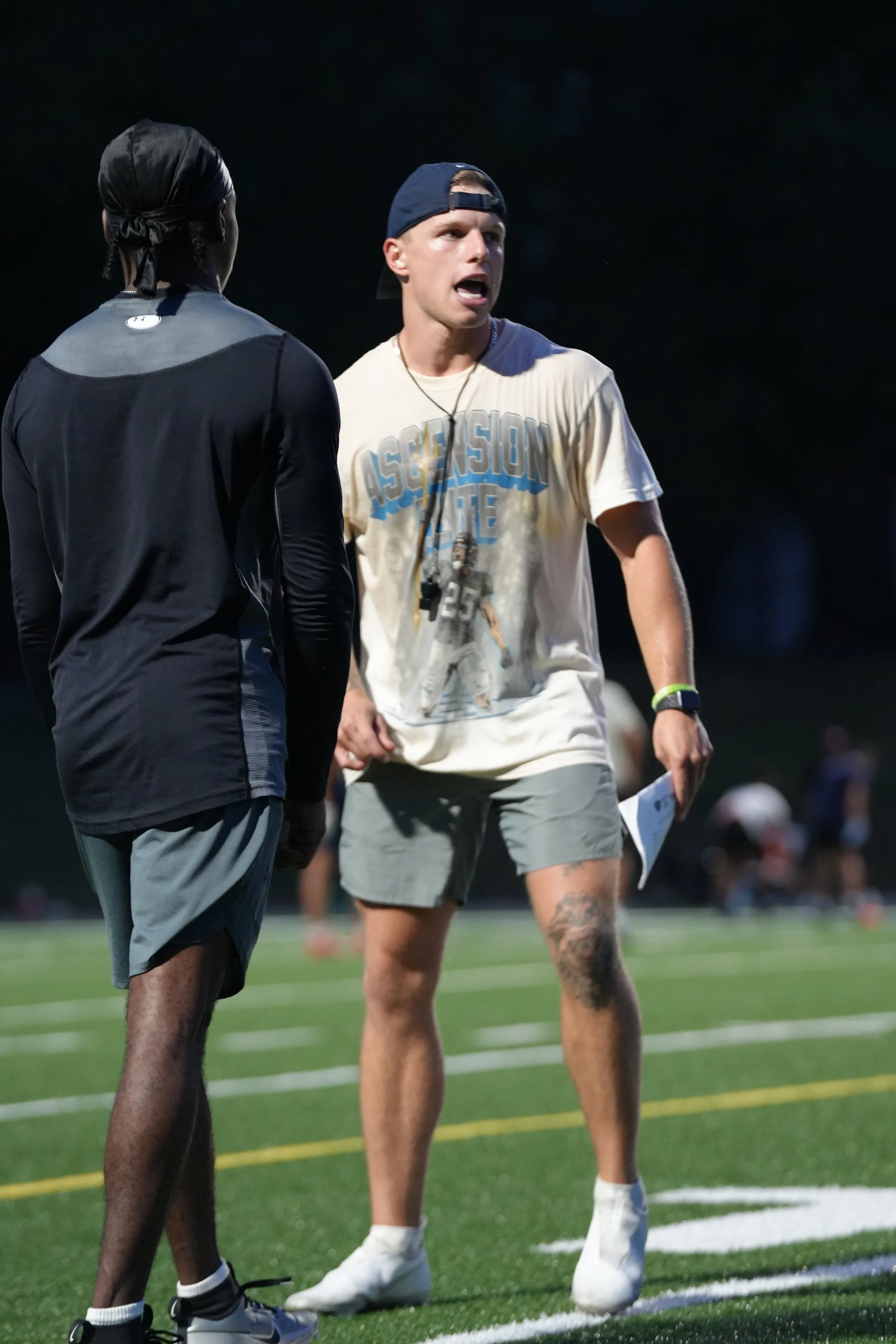 Two young men having a conversation on a football field at night, one wearing a black athletic jacket and shorts, the other in a white t-shirt with graphic print, gray shorts, and a backward baseball cap.