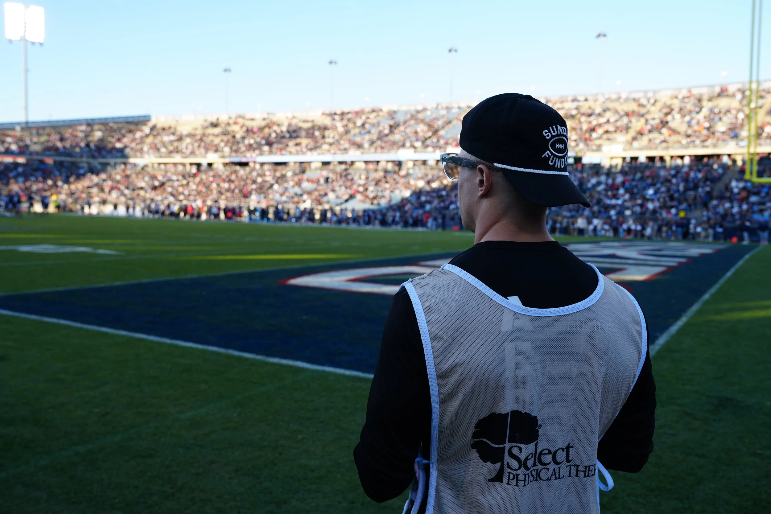A football player or staff member standing on the sidelines of a football field at a stadium, wearing a cap, glasses, and a vest with the logo 'Select PHYSICAL THERAPY'. The stadium is filled with spectators, and the field is green with yard lines and markings.