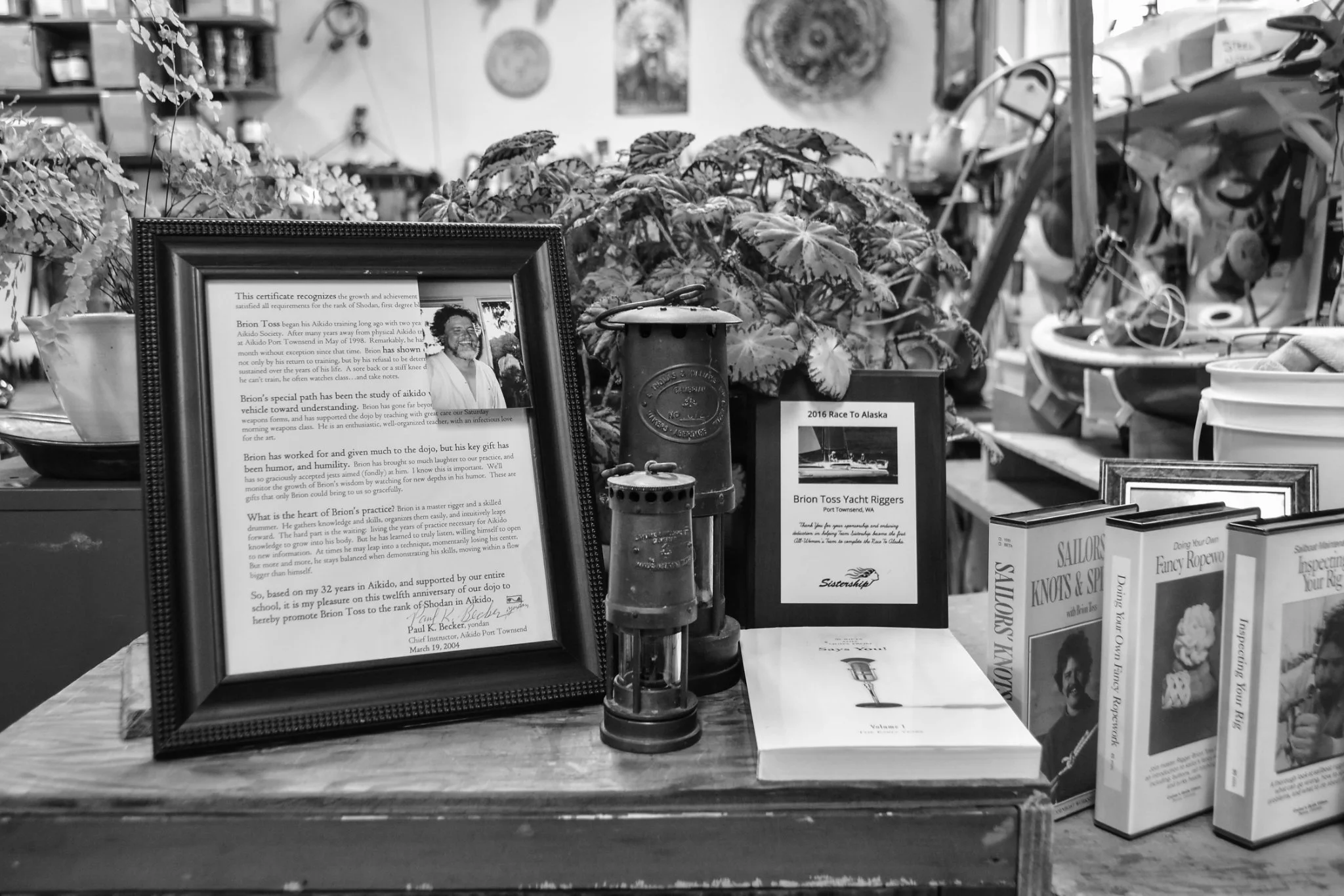 A collection of framed documents, books, and decorative items on a table, with plants and shelves in the background. The main framed document appears to be a certificate of achievement for Brion Toss, and there is a smaller sign related to a yacht rigging event.