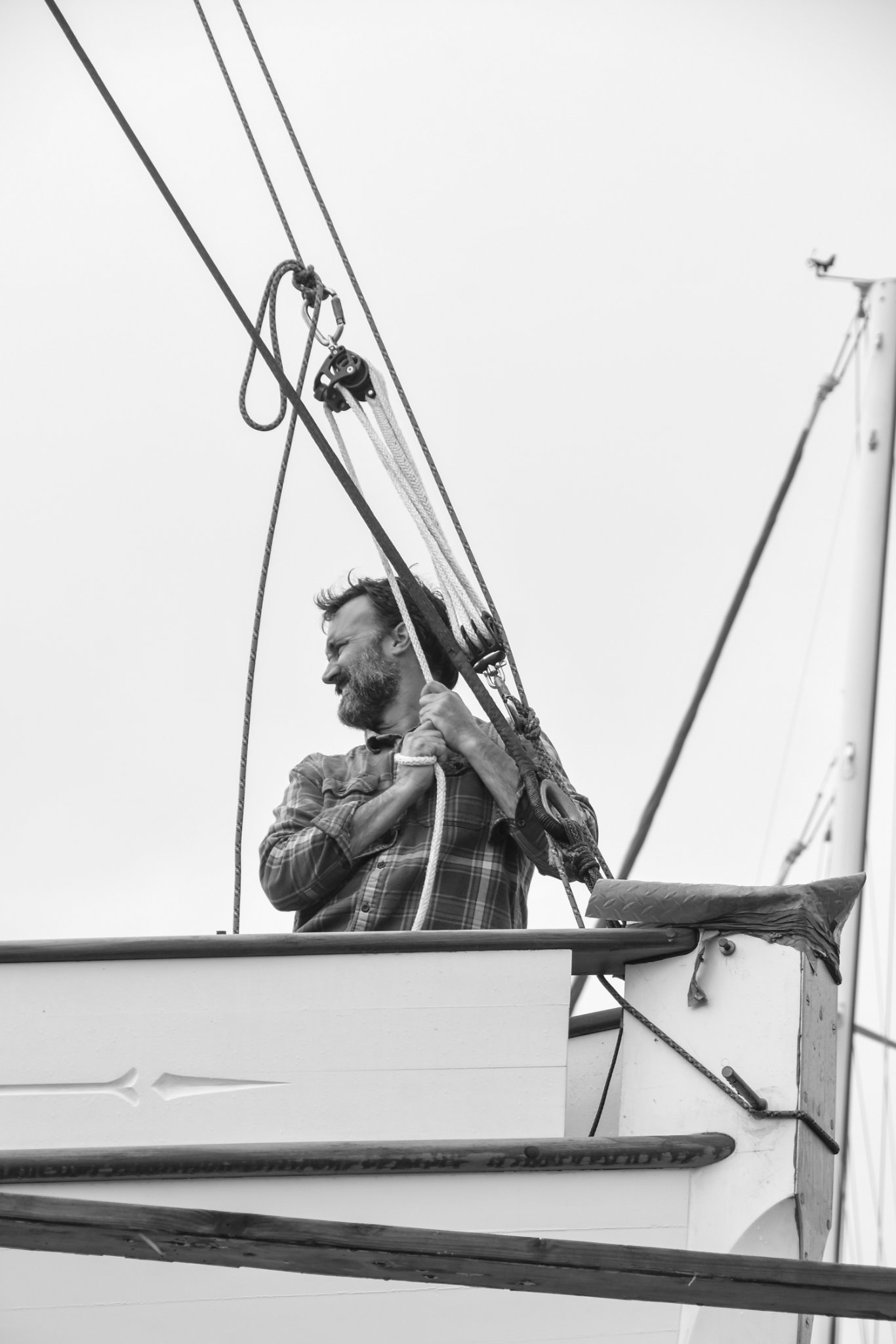 A man with a beard and plaid shirt is on a sailboat, holding lines and smiling. The image is in black and white, showing part of the boat's rigging and sail.
