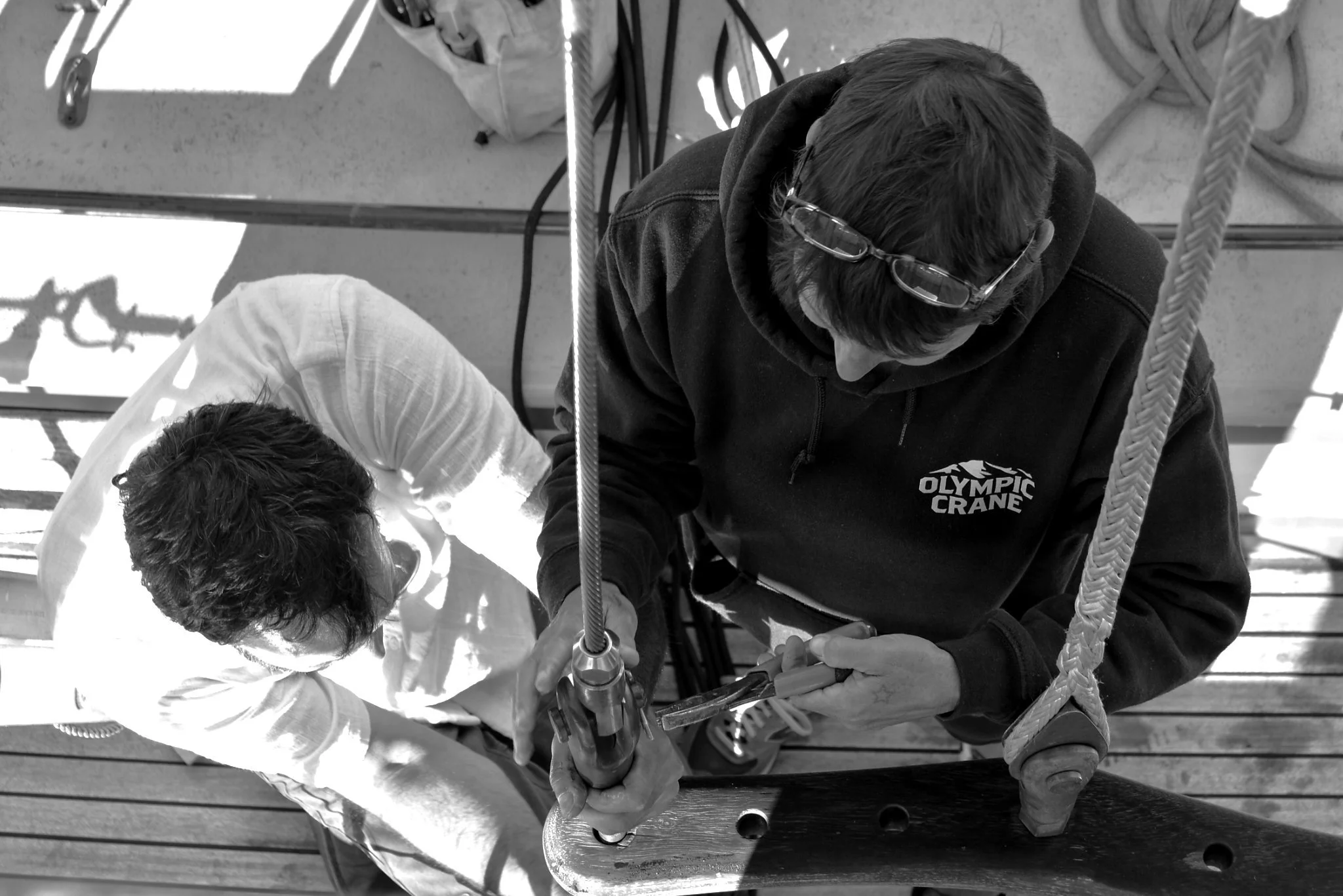 Two individuals working together on new yacht rigging, with tools and supplies nearby; one person in a black hoodie with glasses atop their head and the other in a white shirt.