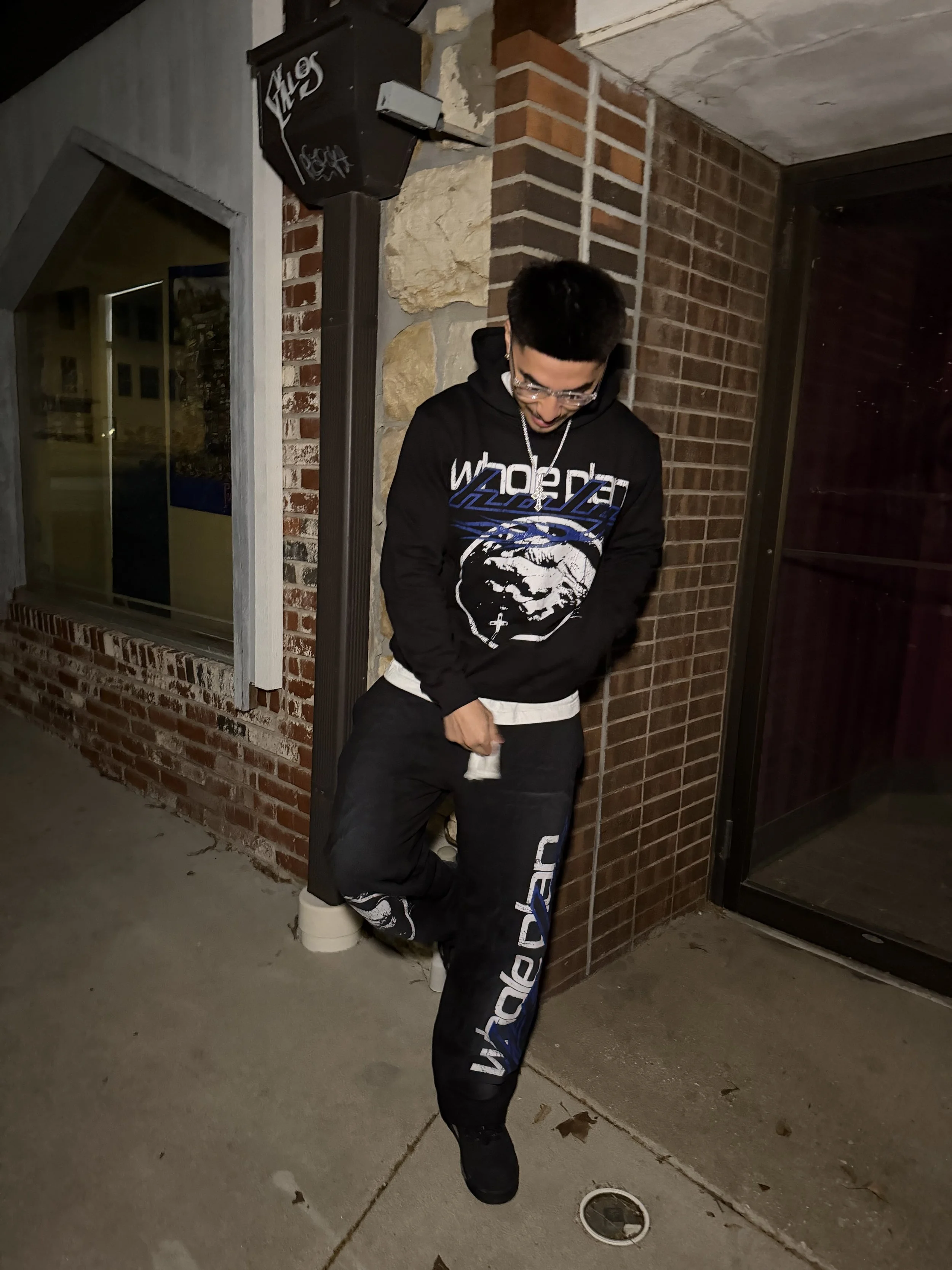A young man with glasses and short dark hair, wearing a black hoodie with graphic print, standing outside at night near a brick wall and window. He is leaning against a corner, looking down, with one foot resting on a small ledge.