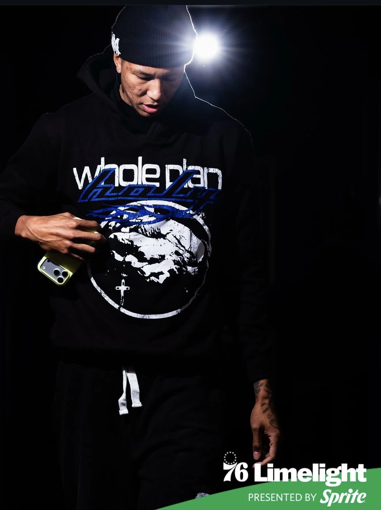 A young man dressed in black outdoor clothing, wearing a black beanie, holding a smartphone, standing in darkness with a bright light behind him, and a logo on the hoodie that reads 'Wade Ryan' with an image of hands in prayer and a cross. The image is part of a promotional poster for the 76th Limelight presented by Sprite.