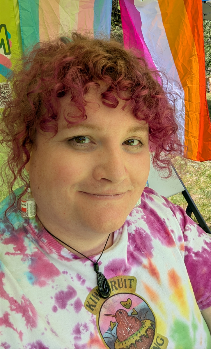 Hazel Bloom, a trans woman with curly hair dyed pink, wearing a rainbow  tie-dye shirt, a pendant necklace, and earrings with estradiol vials. She is smiling in front of lesbian and trans pride flags