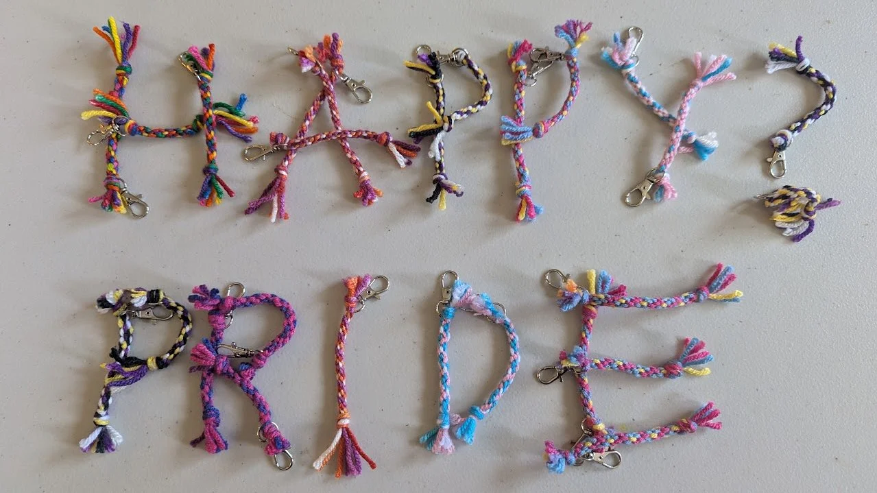 Keychains in the colors of the Lesbian, non-binary, pansexual, bisexual, transgender and rainbow pride flags; they are laid out to spell: "happy? pride"