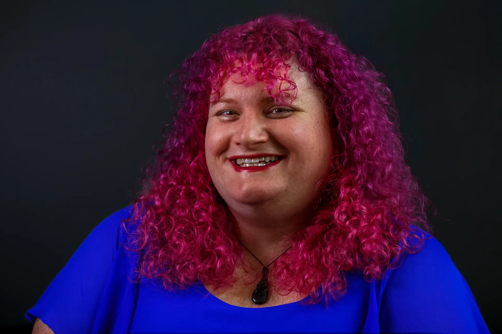 Hazel Bloom, a transgender woman and life coach, smiling, wearing a blue dress and a greenstone necklace, with pink/purple curly hair