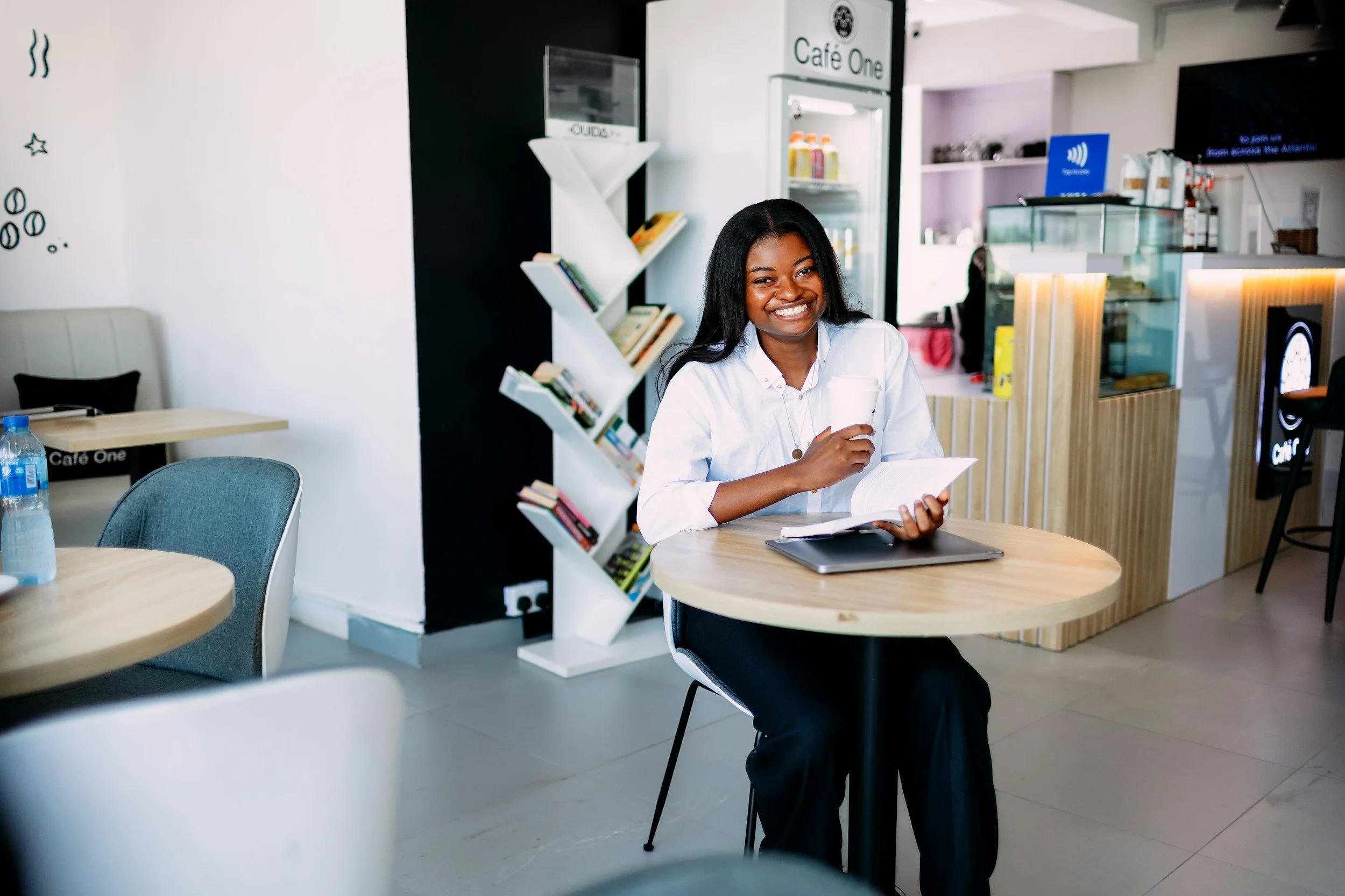 A smiling woman sitting at a round table in a cafe, holding a coffee cup and reading a book, with a laptop on the table.