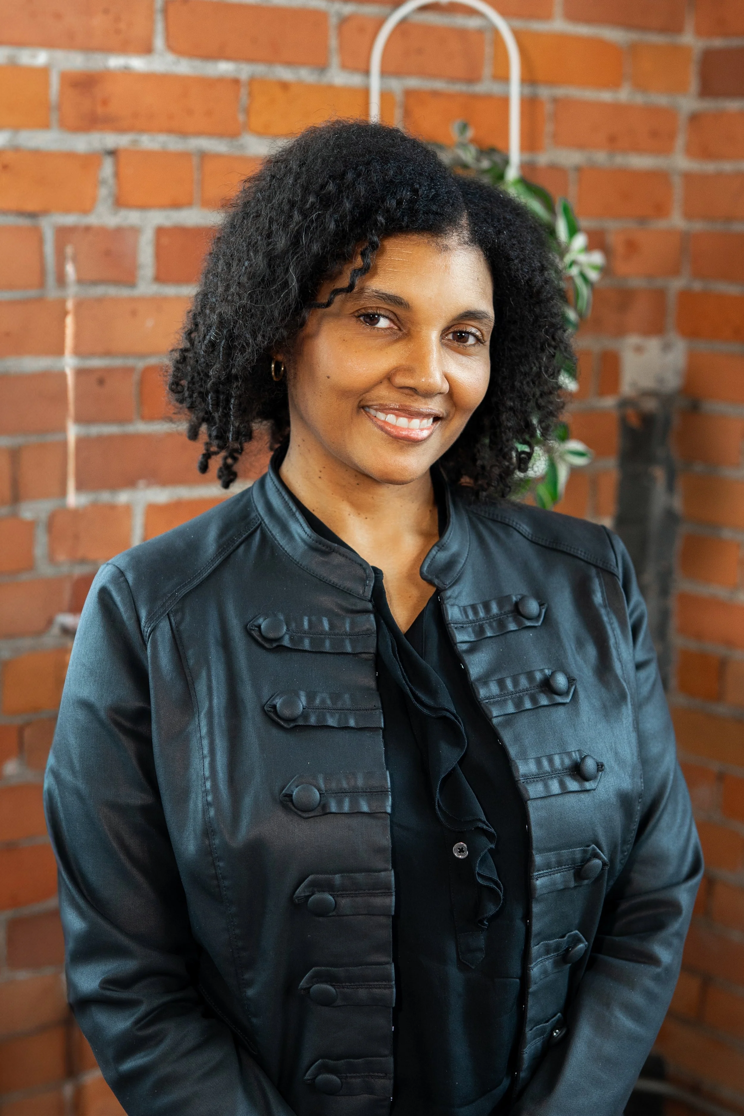 A woman with curly black hair smiling, wearing a black leather jacket, standing in front of a brick wall with a plant in the background.