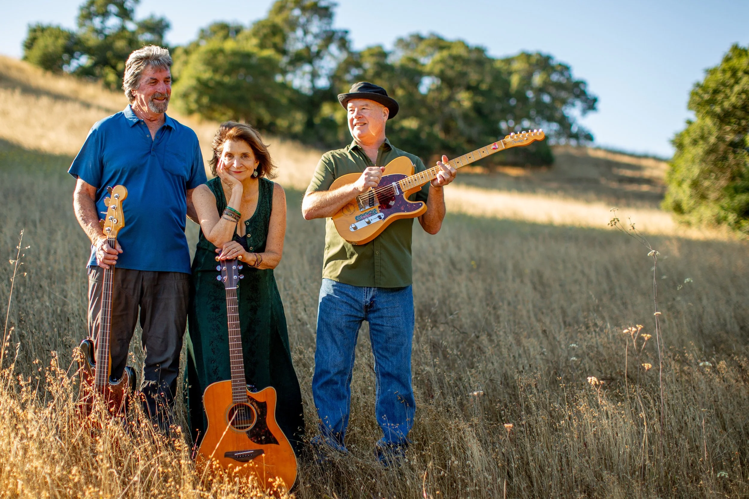 Three musicians standing outdoors in a field with trees in the background, each holding a guitar, with one playing the guitar, on a sunny day.