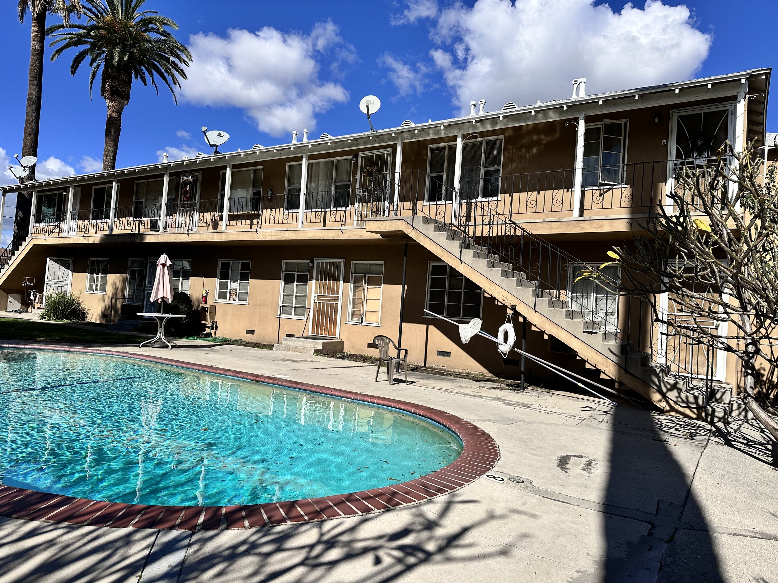 Two-story apartment building with outdoor staircase, a swimming pool in the foreground, palm trees, and a blue sky with clouds.