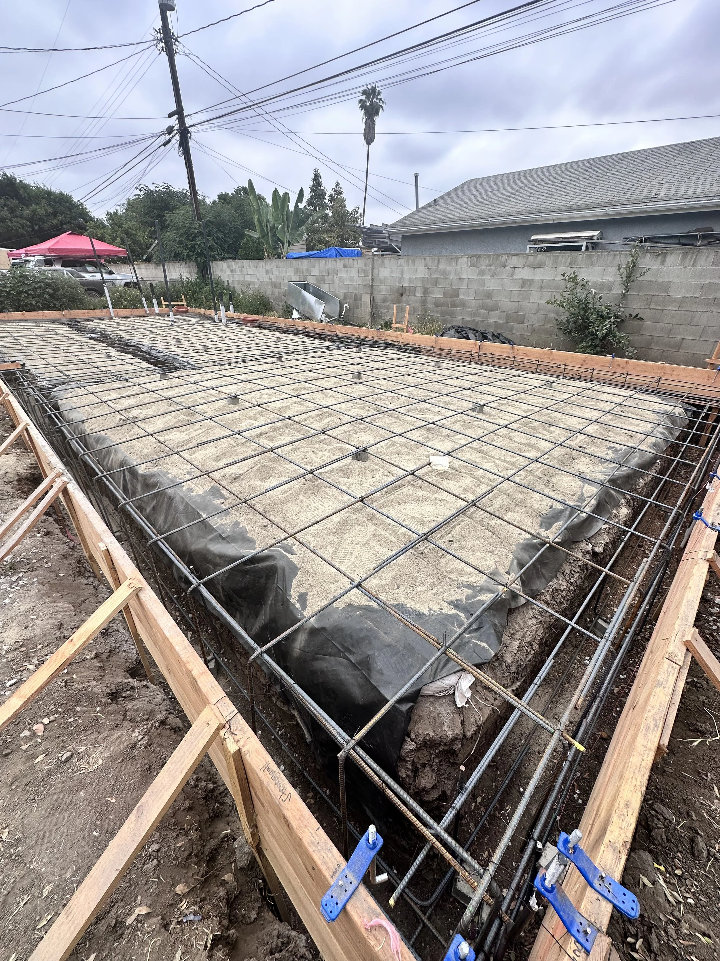 Construction site with rebar and foundation for a building, surrounded by wooden formwork and black plastic sheeting, in a residential backyard.