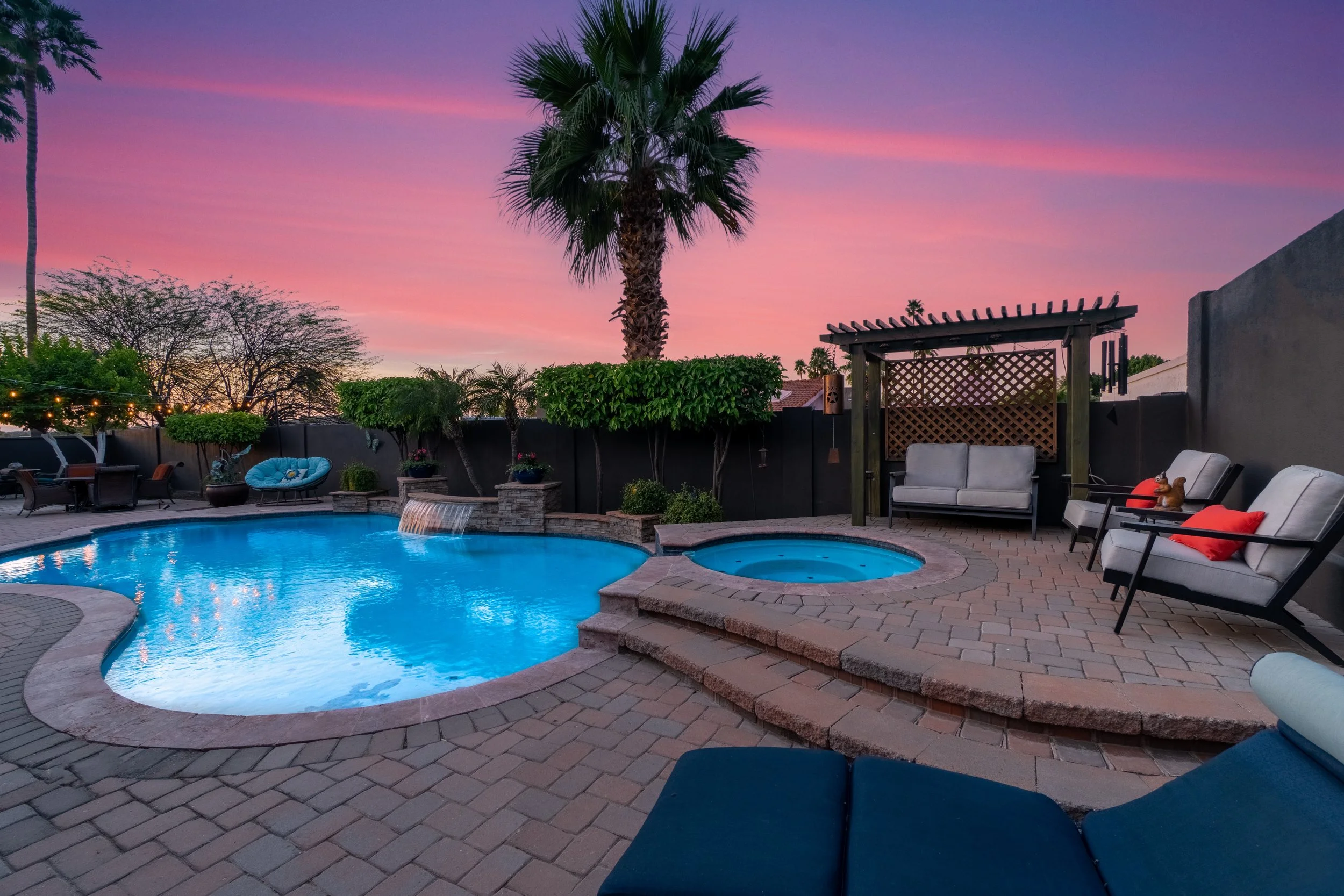 A backyard pool area during sunset with a large palm tree, lounge chairs, a covered swing, and a small hot tub surrounded by brick pavers and lush greenery.