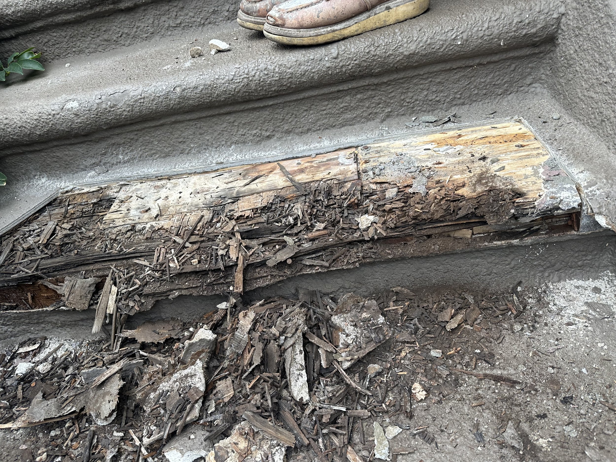 Close-up of a damaged wooden stair tread with broken and splintered wood, debris, and dirt, with a pair of sandals visible at the top of the stairs.