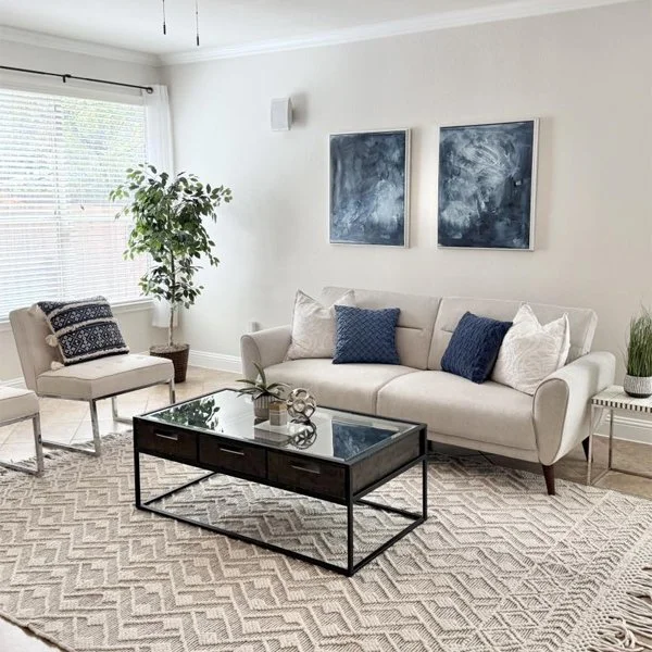 Modern living room with a beige sofa, two blue abstract paintings, a glass coffee table, and a patterned rug.
