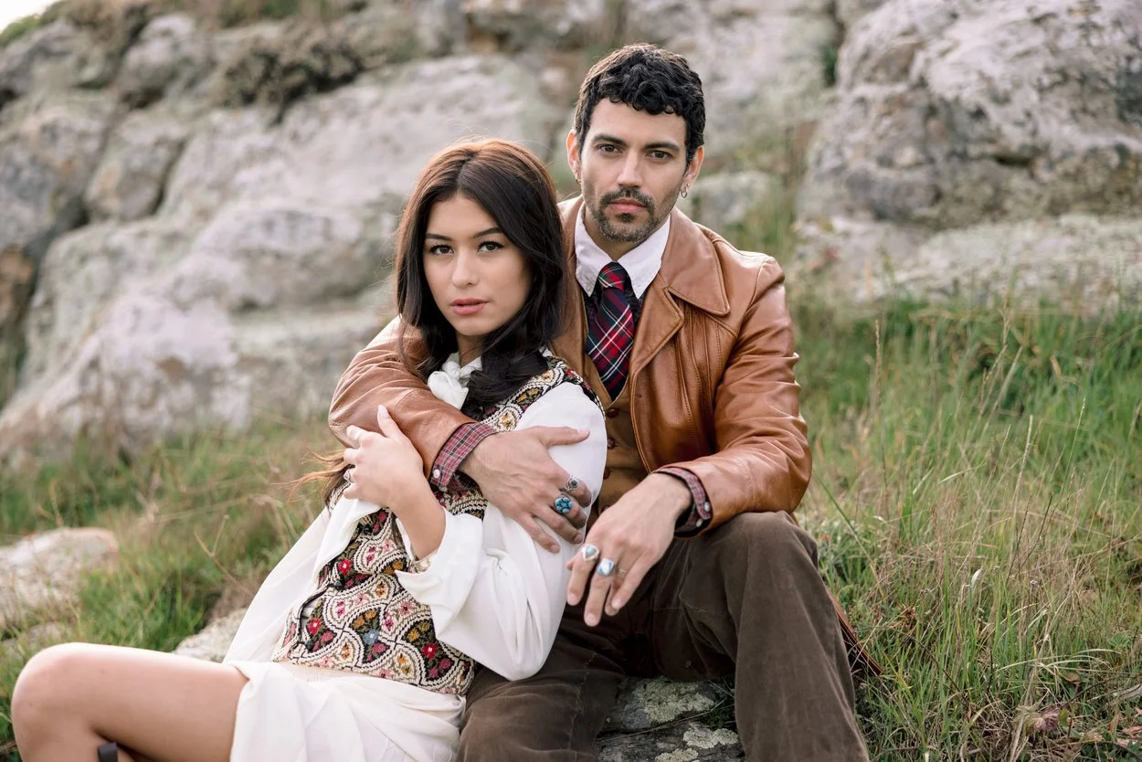 A man and woman sitting outdoors on grass near rocks, with the man embracing the woman. The woman has long dark hair and is wearing a white dress with an embroidered vest. The man has curly dark hair, a beard, and is wearing a brown leather jacket over a red plaid shirt and tie.