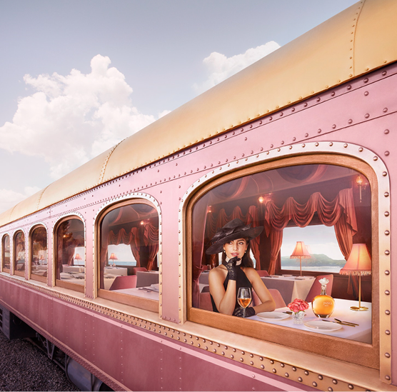 A woman in vintage attire, including a black hat and gloves, sitting at a window table inside a pink dining train car with a glass of wine, flowers, and a lamp, outside view shows a cloudy sky.