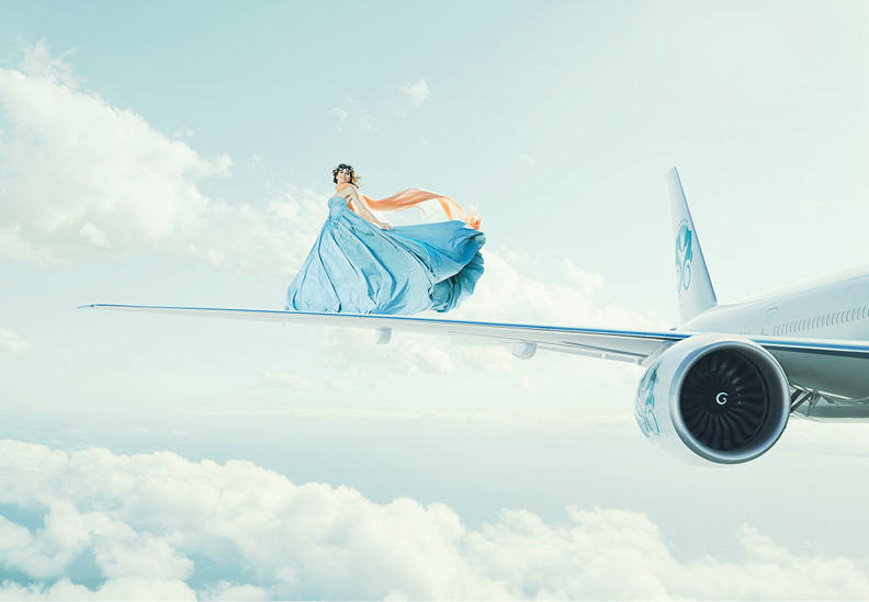 A woman in a flowing blue dress and orange scarf is standing on the wing of a commercial airplane flying high above the clouds in the sky.