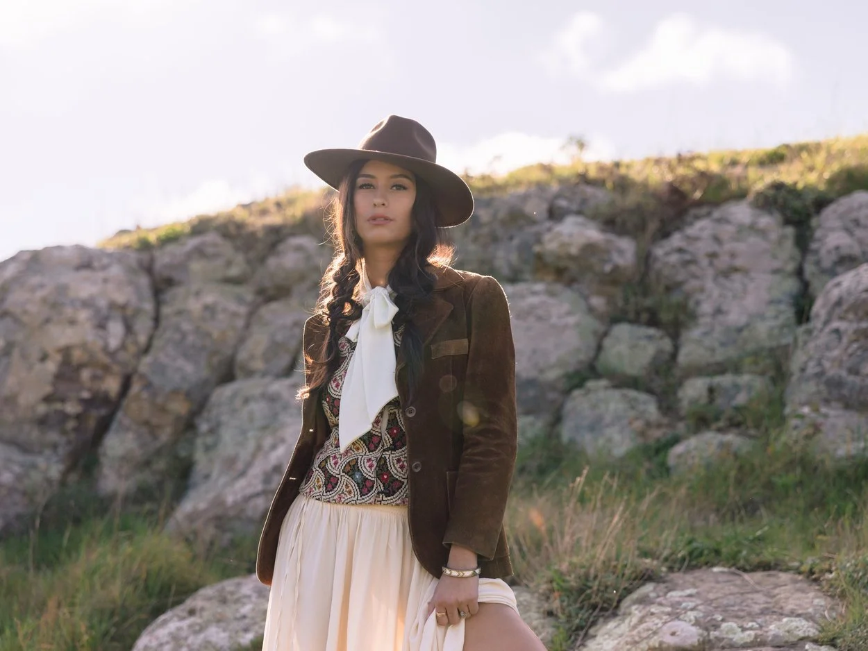 A woman standing outdoors in front of rocks, wearing a wide-brimmed hat, a patterned vest, a white shirt with a bow tie, and a beige skirt.