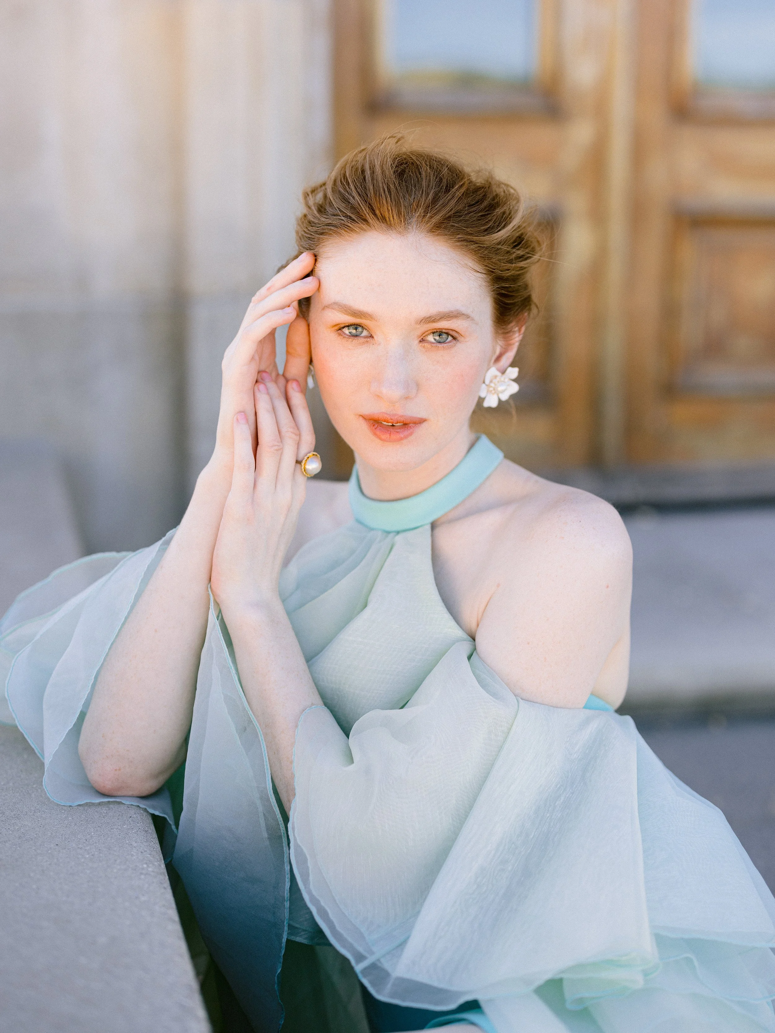 A woman with red hair and blue eyes wearing a light blue dress and matching choker, posing outdoors with her hand on her face, near a wooden door.